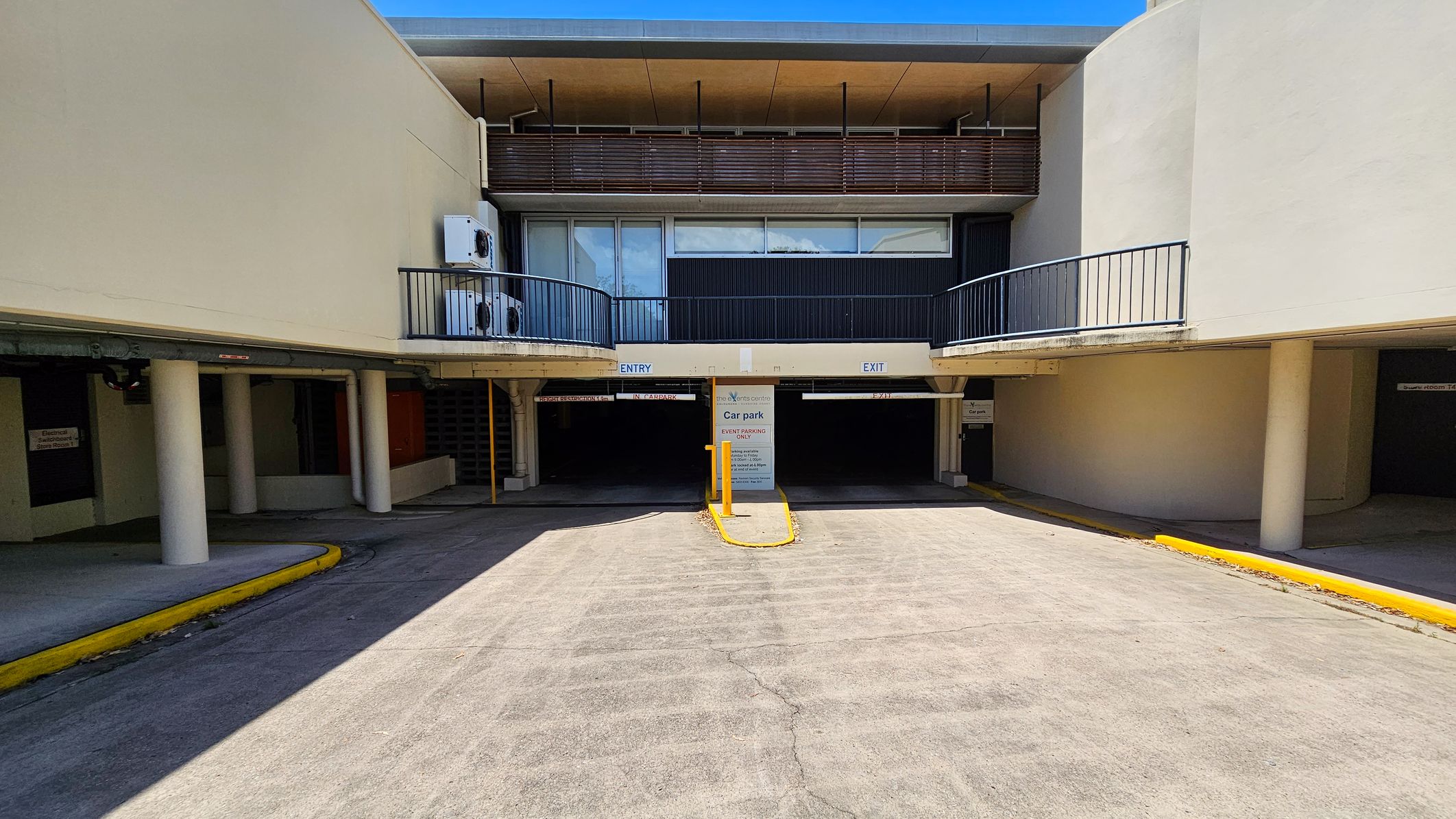 On the left, a curved road with parked vehicles and a yellow line along the edge. A large tree is visible near the centre. On the right, a building with a flat roof and a balcony, featuring air conditioning units. Below, a car park with a sign indicating "Café Open" and another sign for event parking. A small orange cone is on the pavement near the road. The ground is a mix of concrete and grass. On the left, a curved road with parked vehicles and a yellow line along the edge. A large tree is visible near the centre. On the right, a building with a flat roof and a balcony, featuring air conditioning units. Below, a car park with a sign indicating "Café Open" and another sign for event parking. A small orange cone is on the pavement near the road. The ground is a mix of concrete and grass.