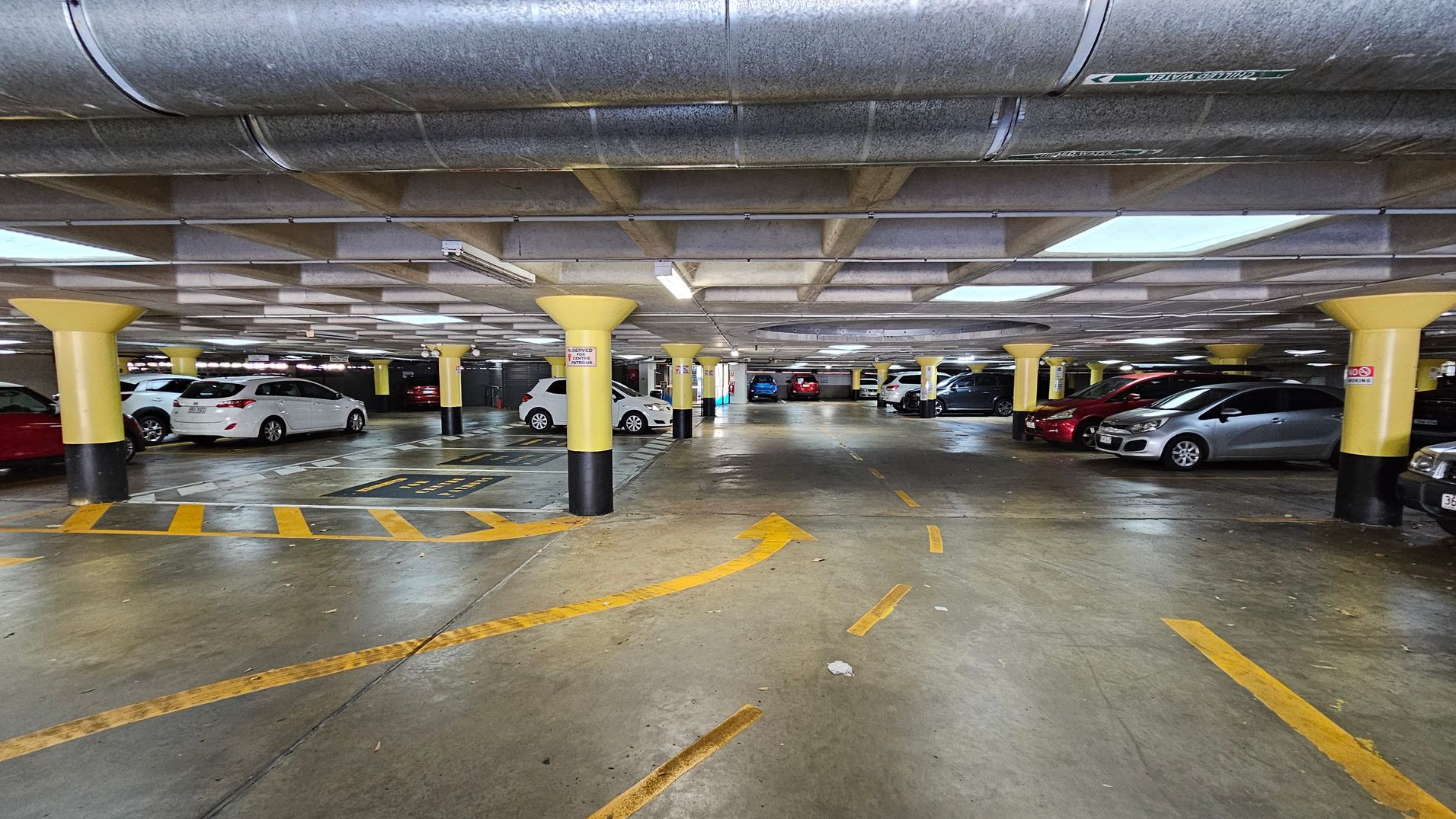 Underground car park with a concrete floor marked with yellow directional arrows and lines. Yellow and black pillars support the low ceiling, which has exposed ductwork and fluorescent lighting. Cars are parked on both sides, including white, red, and silver vehicles. The area is well-lit, and the space appears clean and organised. Underground car park with a concrete floor marked with yellow directional arrows and lines. Yellow and black pillars support the low ceiling, which has exposed ductwork and fluorescent lighting. Cars are parked on both sides, including white, red, and silver vehicles. The area is well-lit, and the space appears clean and organised.