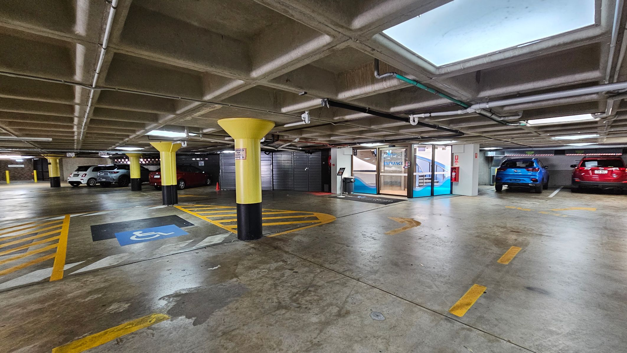 Underground car park with a concrete floor and ceiling. On the left, several cars are parked, including a white and a silver vehicle. In the centre, a yellow column stands near a marked disabled parking space with blue and yellow lines. On the right, a blue and a red car are parked. The background features a glass entrance door with a sign above it. The ceiling has visible pipes and rectangular skylights. Underground car park with a concrete floor and ceiling. On the left, several cars are parked, including a white and a silver vehicle. In the centre, a yellow column stands near a marked disabled parking space with blue and yellow lines. On the right, a blue and a red car are parked. The background features a glass entrance door with a sign above it. The ceiling has visible pipes and rectangular skylights.