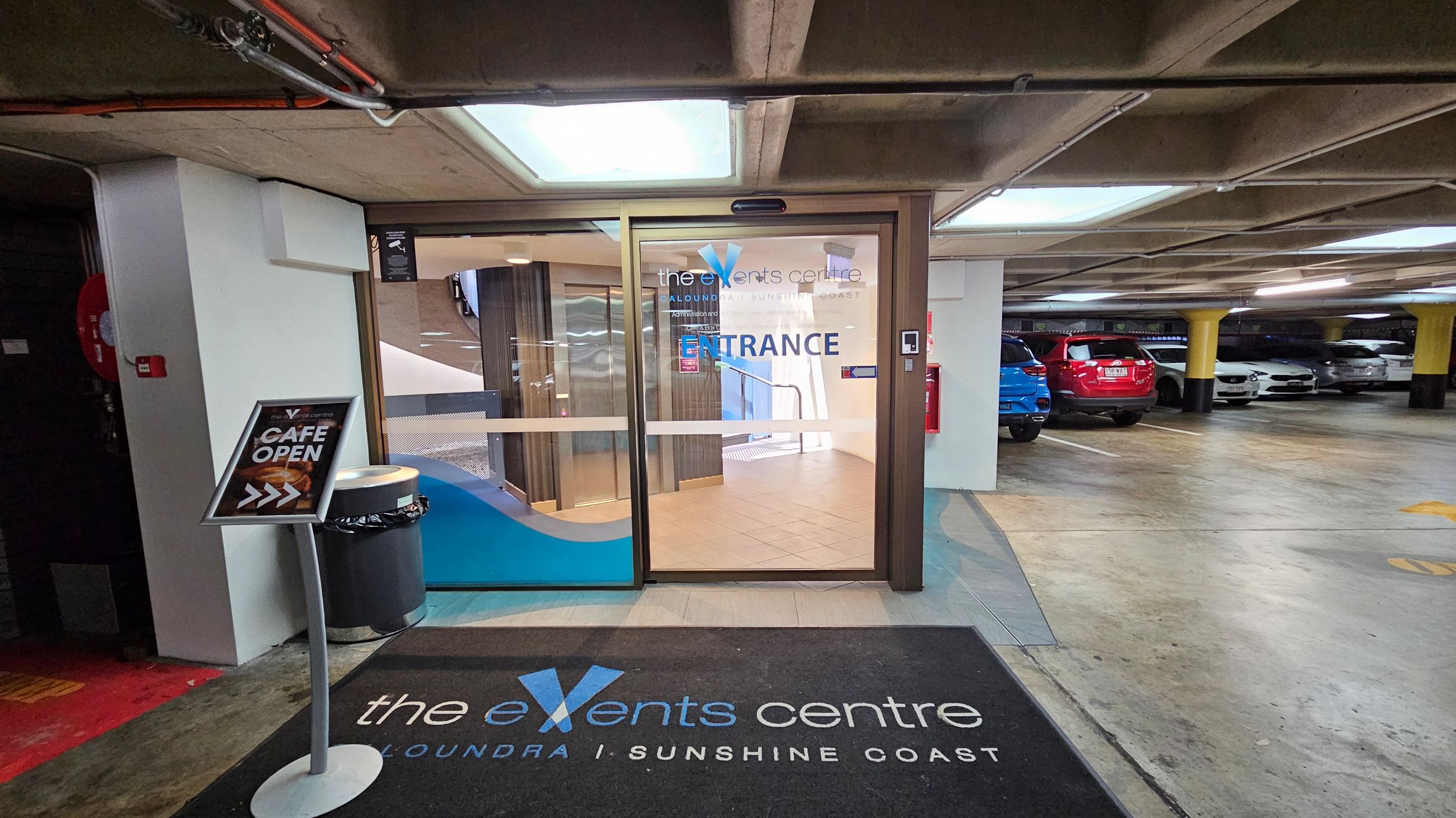 Entrance to a venue from a car park. On the left, a sign reads "CAFE OPEN" with an arrow, next to a bin. The glass door in the centre has "the events centre" and "ENTRANCE" written on it. Inside, a staircase is visible. On the right, several parked cars, including a red and a blue one, are in the car park. The floor is concrete, and the ceiling has exposed pipes and lights. A black mat with "the events centre" is on the floor in front of the door. Entrance to a venue from a car park. On the left, a sign reads "CAFE OPEN" with an arrow, next to a bin. The glass door in the centre has "the events centre" and "ENTRANCE" written on it. Inside, a staircase is visible. On the right, several parked cars, including a red and a blue one, are in the car park. The floor is concrete, and the ceiling has exposed pipes and lights. A black mat with "the events centre" is on the floor in front of the door.