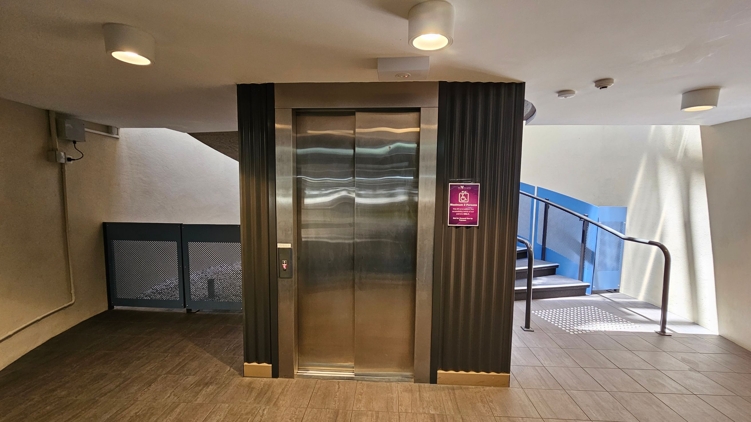 A modern lift with shiny metallic doors is centred in the image, flanked by dark vertical panels. To the right, a staircase with a blue railing ascends, with a textured mat at the base. On the left, a wall-mounted electrical box and a vent are visible. The floor is tiled in a light brown shade, and the ceiling is white with recessed lighting fixtures. A sign with a pink background is mounted next to the lift. A modern lift with shiny metallic doors is centred in the image, flanked by dark vertical panels. To the right, a staircase with a blue railing ascends, with a textured mat at the base. On the left, a wall-mounted electrical box and a vent are visible. The floor is tiled in a light brown shade, and the ceiling is white with recessed lighting fixtures. A sign with a pink background is mounted next to the lift.