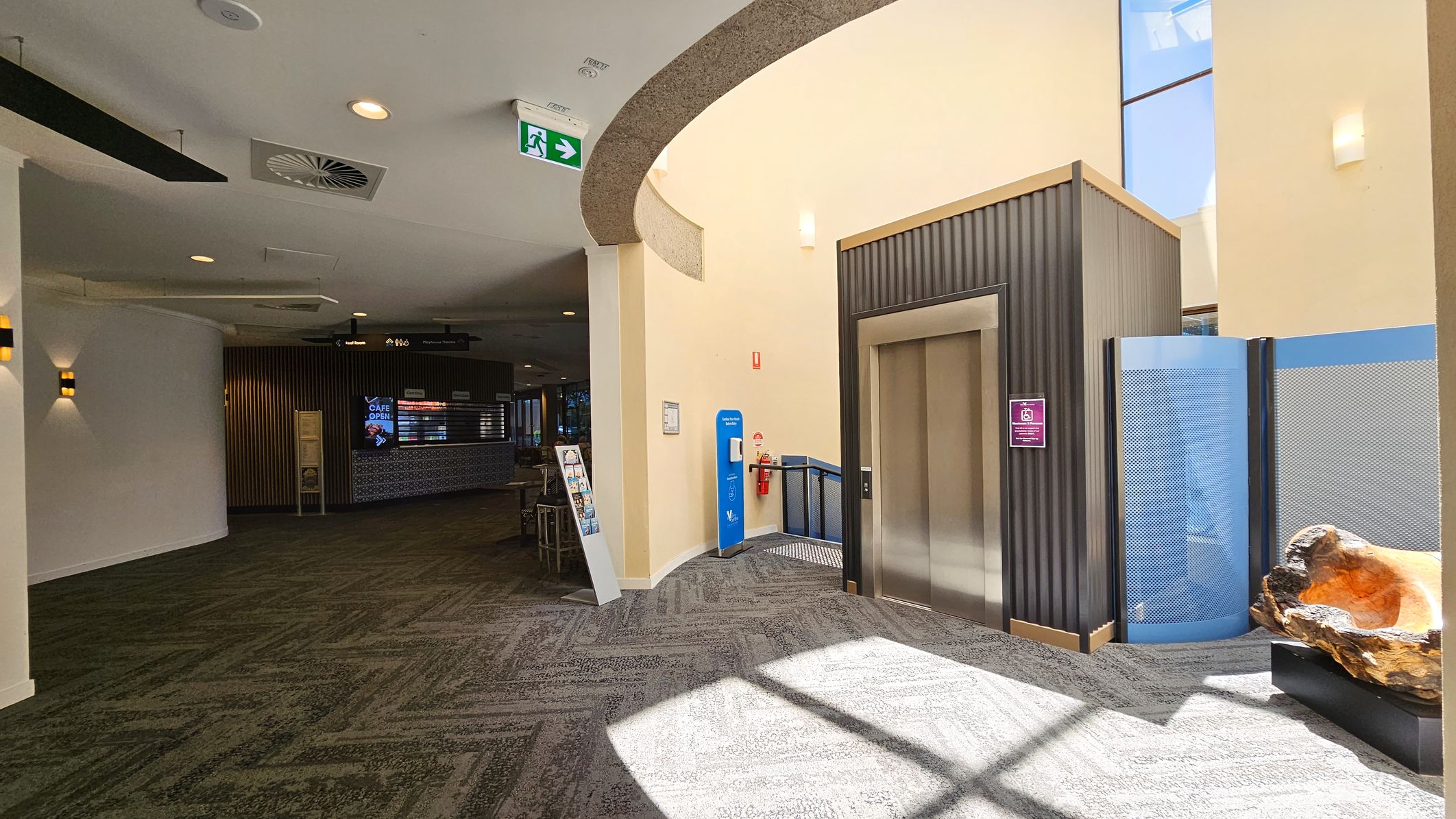 On the left, a curved wall with wall-mounted lights and a green carpeted floor. In the middle, an elevator with a metallic door, a blue hand sanitiser station, and a fire extinguisher. On the right, a decorative wooden sculpture on a black stand and a blue partition. The ceiling is light-coloured with recessed lighting and ventilation. An exit sign is visible above. In the background, a café area with a digital menu board. On the left, a curved wall with wall-mounted lights and a green carpeted floor. In the middle, an elevator with a metallic door, a blue hand sanitiser station, and a fire extinguisher. On the right, a decorative wooden sculpture on a black stand and a blue partition. The ceiling is light-coloured with recessed lighting and ventilation. An exit sign is visible above. In the background, a café area with a digital menu board.
