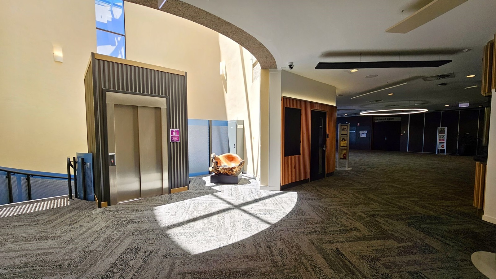 A modern interior with a lift on the left, featuring a metallic door and a striped panel design. To the right, a wooden panelled wall with a dark doorway. In the centre, a large, polished wooden sculpture sits on a pedestal, illuminated by natural light from above. The floor is carpeted in a green and grey pattern, and the ceiling has recessed lighting and a circular light fixture in the background. A modern interior with a lift on the left, featuring a metallic door and a striped panel design. To the right, a wooden panelled wall with a dark doorway. In the centre, a large, polished wooden sculpture sits on a pedestal, illuminated by natural light from above. The floor is carpeted in a green and grey pattern, and the ceiling has recessed lighting and a circular light fixture in the background.