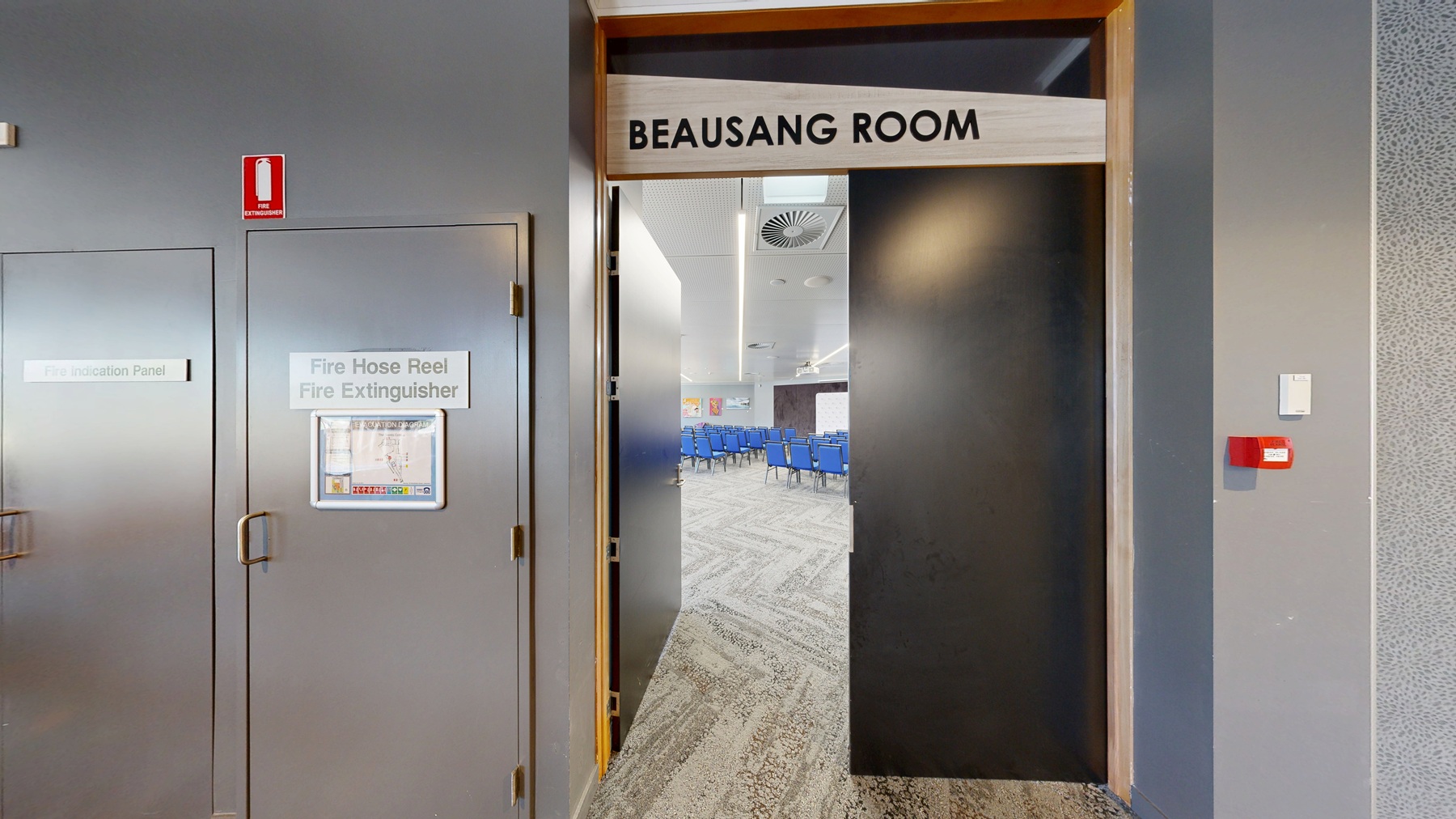 On the left, a room with rows of blue chairs facing forward, with colourful artwork on the walls. The floor is carpeted in a light grey pattern. On the right, a hallway with a brown and green patterned carpet, featuring several large potted plants. A digital screen is mounted on the wall displaying an advertisement. The ceiling has recessed lighting and air vents. A partially open door with a sign above it reads "ROOM". On the left, a room with rows of blue chairs facing forward, with colourful artwork on the walls. The floor is carpeted in a light grey pattern. On the right, a hallway with a brown and green patterned carpet, featuring several large potted plants. A digital screen is mounted on the wall displaying an advertisement. The ceiling has recessed lighting and air vents. A partially open door with a sign above it reads "ROOM".