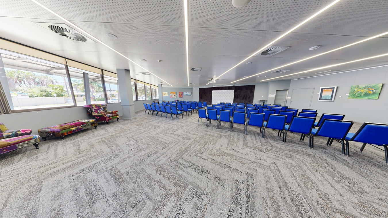 A spacious conference room with a grey patterned carpet and a white ceiling featuring linear lights. On the right, rows of blue chairs are arranged facing a white screen at the front. The left side has colourful, patterned sofas near large windows that let in natural light. The walls are adorned with framed artwork. A spacious conference room with a grey patterned carpet and a white ceiling featuring linear lights. On the right, rows of blue chairs are arranged facing a white screen at the front. The left side has colourful, patterned sofas near large windows that let in natural light. The walls are adorned with framed artwork.