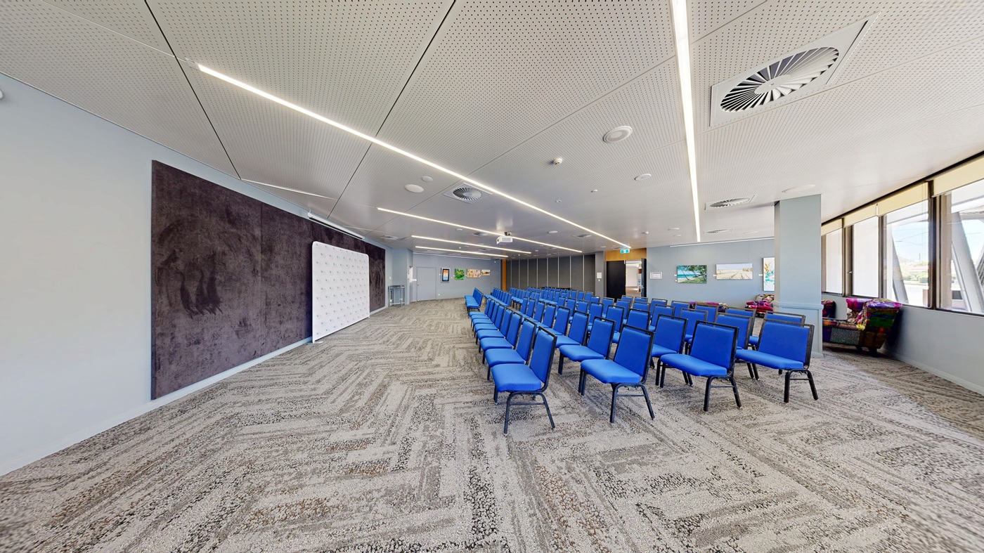 A conference room with rows of blue chairs arranged in the centre. On the left, there's a large dark panel on the wall and a white board. The right side features large windows with blinds partially drawn, allowing natural light in. The floor is covered with a patterned carpet in shades of grey and green. The ceiling is white with recessed lighting and air vents. In the background, there are framed pictures on the wall and a small seating area with colourful cushions. A conference room with rows of blue chairs arranged in the centre. On the left, there's a large dark panel on the wall and a white board. The right side features large windows with blinds partially drawn, allowing natural light in. The floor is covered with a patterned carpet in shades of grey and green. The ceiling is white with recessed lighting and air vents. In the background, there are framed pictures on the wall and a small seating area with colourful cushions.