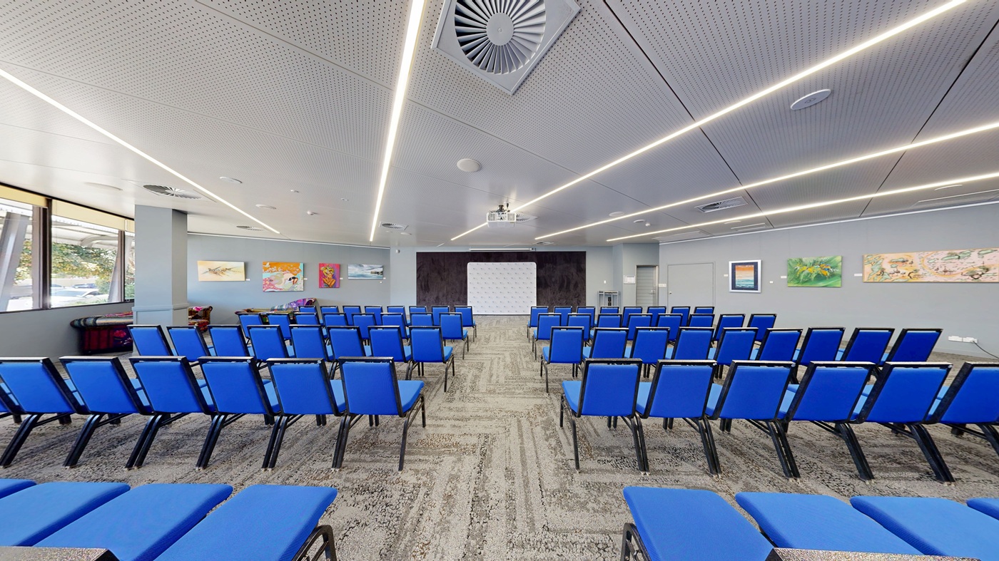 A conference room with rows of blue chairs facing a white projection screen at the front. The walls are adorned with colourful artwork. The ceiling features linear lights and a large air vent. The floor is carpeted with a patterned design. Windows on the left allow natural light to enter, and a small table with items is visible near the windows. A conference room with rows of blue chairs facing a white projection screen at the front. The walls are adorned with colourful artwork. The ceiling features linear lights and a large air vent. The floor is carpeted with a patterned design. Windows on the left allow natural light to enter, and a small table with items is visible near the windows.