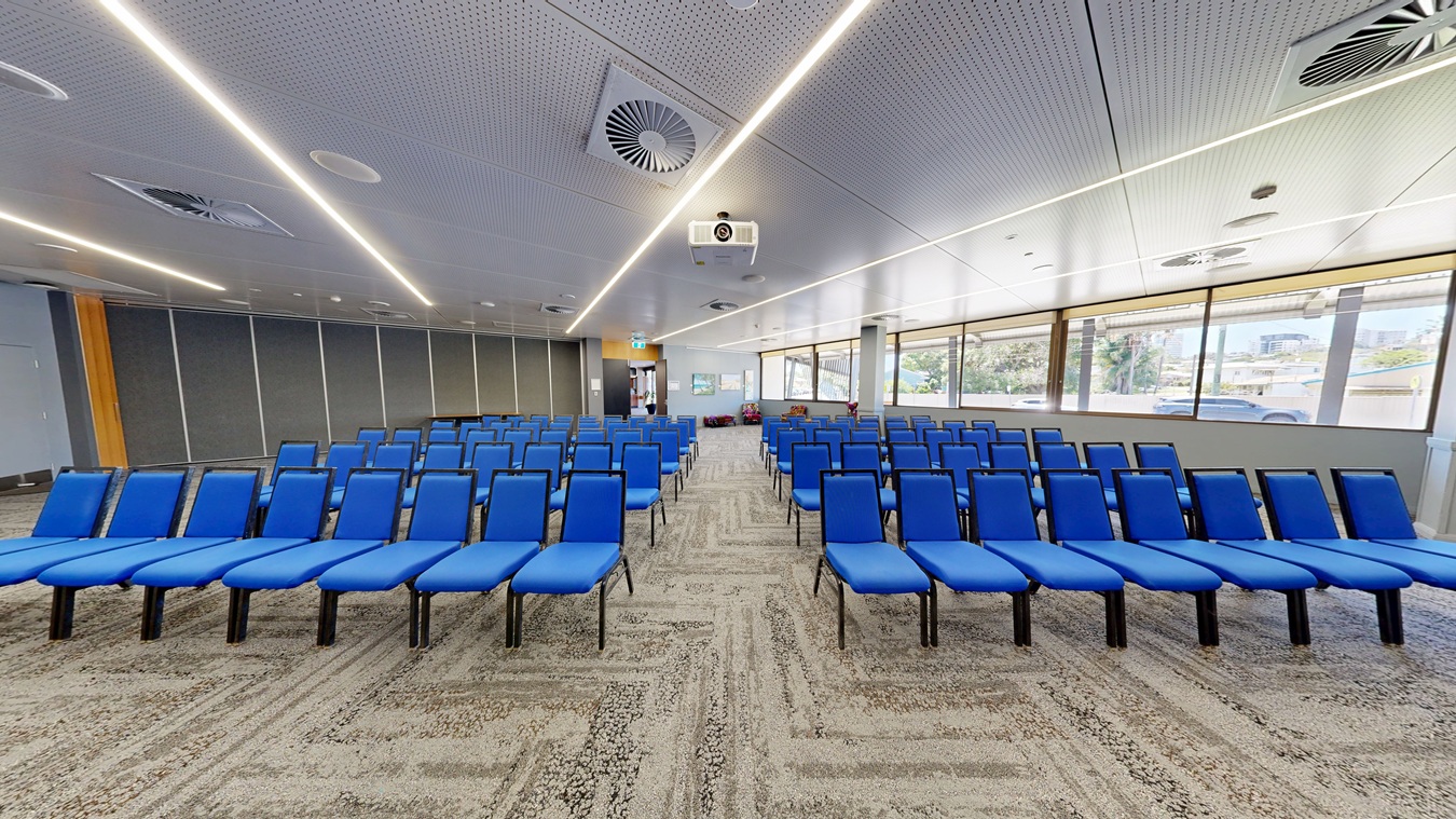 Rows of blue chairs are arranged in a conference room. On the left, there are grey partition walls. On the right, large windows allow natural light to enter, showing a view of trees and buildings outside. The ceiling is white with recessed lighting and air vents. The floor is carpeted in a patterned design. At the back, there is a projector mounted on the ceiling and an exit sign above a door. Rows of blue chairs are arranged in a conference room. On the left, there are grey partition walls. On the right, large windows allow natural light to enter, showing a view of trees and buildings outside. The ceiling is white with recessed lighting and air vents. The floor is carpeted in a patterned design. At the back, there is a projector mounted on the ceiling and an exit sign above a door.