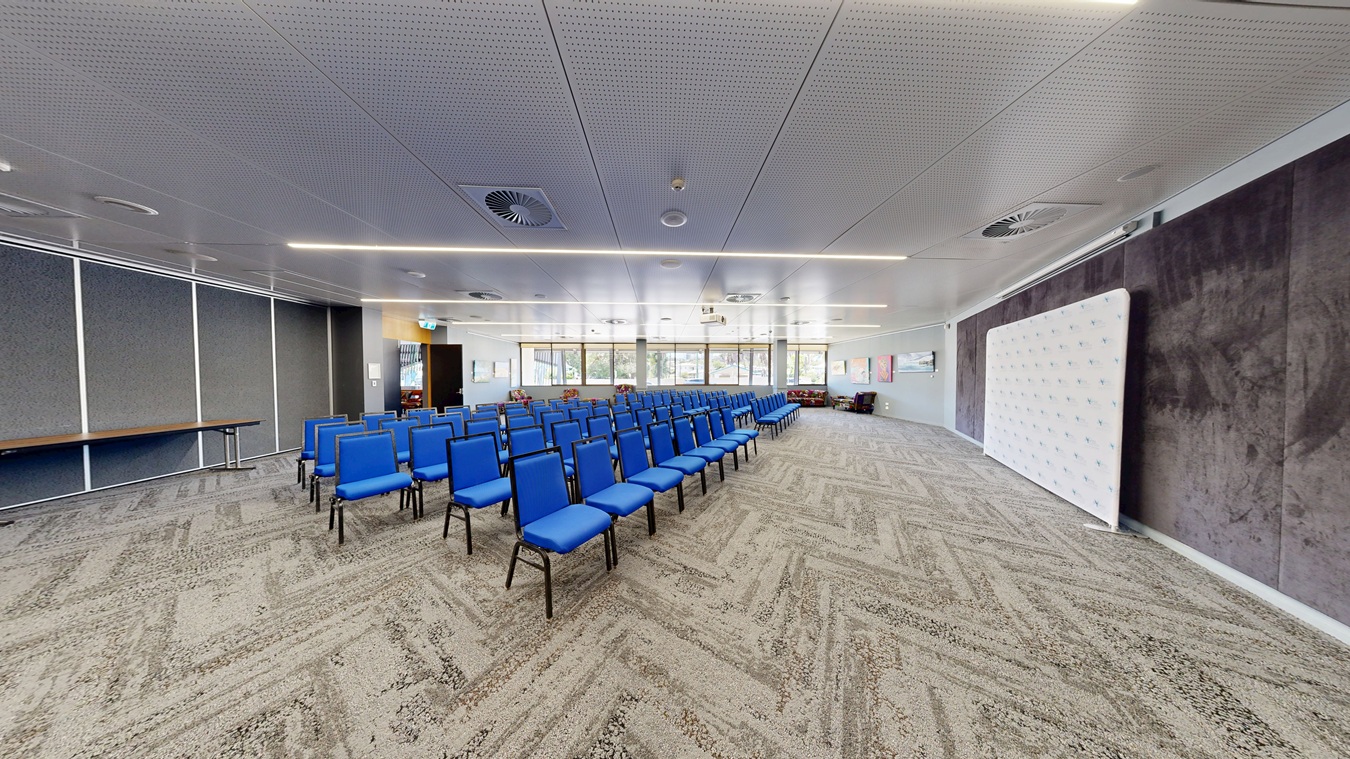 A conference room with rows of blue chairs arranged in the centre. On the left, there is a long table against a wall with vertical panels. The right side features a large white display board. The background has large windows allowing natural light, and the floor is covered with a patterned carpet. The ceiling is white with recessed lighting and circular air vents. A conference room with rows of blue chairs arranged in the centre. On the left, there is a long table against a wall with vertical panels. The right side features a large white display board. The background has large windows allowing natural light, and the floor is covered with a patterned carpet. The ceiling is white with recessed lighting and circular air vents.
