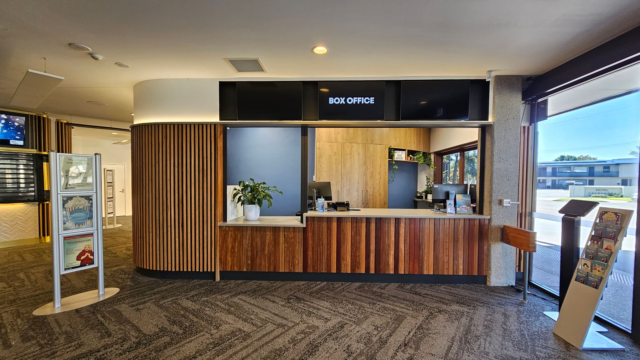 Entrance to a building with large glass doors. On the left, a reflection shows palm trees and a street. The right side features wooden panelling with part of the word "Box" visible. Inside, a carpeted area with a circular light fixture on the ceiling, a reception desk, and digital screens. A black mat at the entrance reads "the events centre CALOUNDRA | SUNSHINE COAST." The floor outside is light-coloured with a textured surface. Entrance to a building with large glass doors. On the left, a reflection shows palm trees and a street. The right side features wooden panelling with part of the word "Box" visible. Inside, a carpeted area with a circular light fixture on the ceiling, a reception desk, and digital screens. A black mat at the entrance reads "the events centre CALOUNDRA | SUNSHINE COAST." The floor outside is light-coloured with a textured surface.
