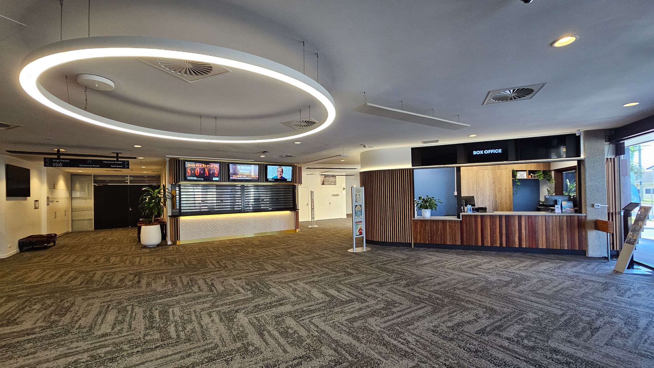 A spacious lobby with a patterned carpet in shades of green and grey. On the right, there's a box office with a wooden counter and a plant. In the centre, a circular light fixture hangs from the ceiling. To the left, a digital display board and a potted plant are visible. The ceiling has recessed lighting and air vents. Signs for different theatre rooms are visible in the background. A spacious lobby with a patterned carpet in shades of green and grey. On the right, there's a box office with a wooden counter and a plant. In the centre, a circular light fixture hangs from the ceiling. To the left, a digital display board and a potted plant are visible. The ceiling has recessed lighting and air vents. Signs for different theatre rooms are visible in the background.