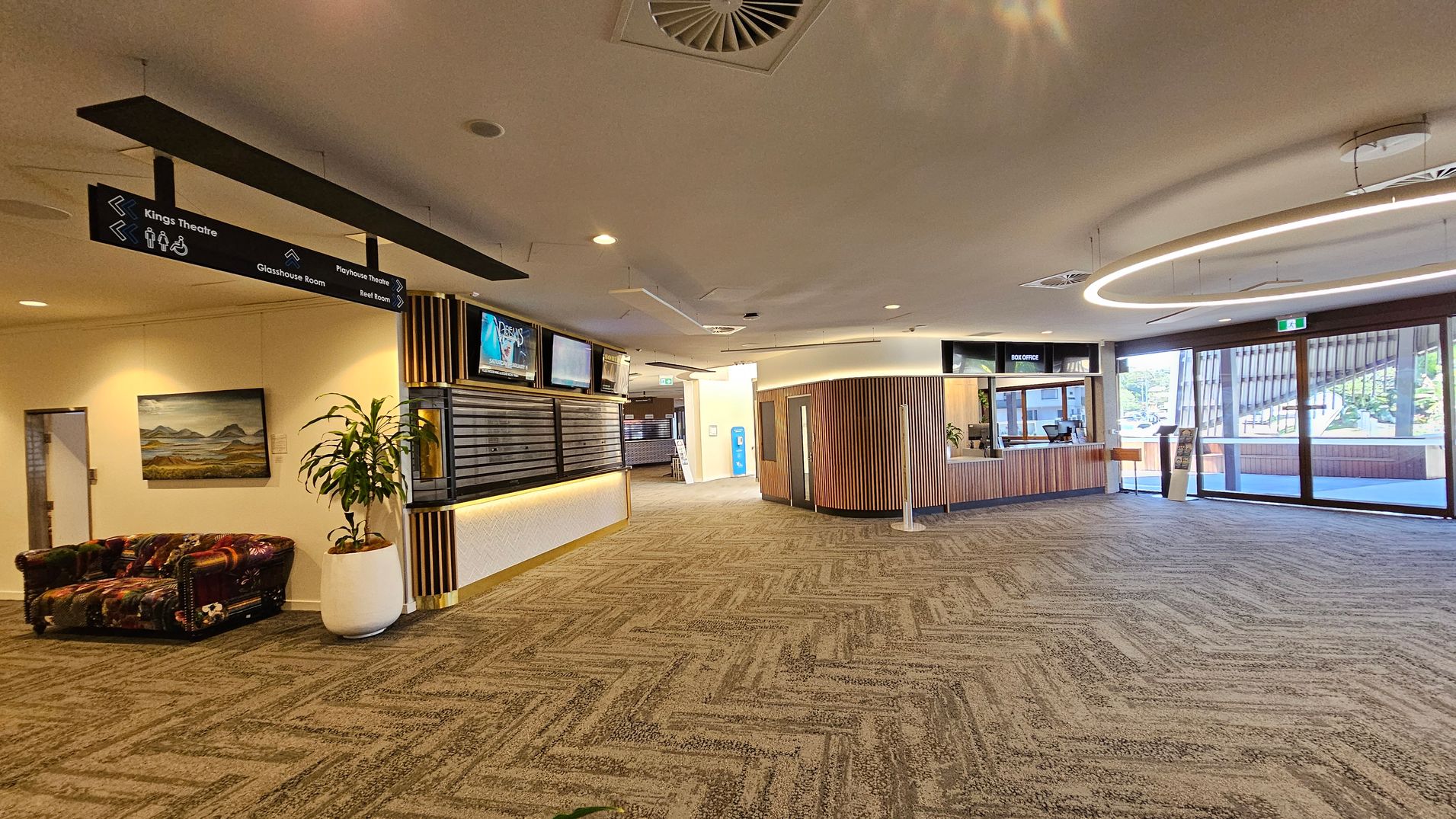 Spacious lobby with patterned carpet flooring. On the left, a colourful sofa and a painting on the wall. Overhead signs direct to various rooms. In the centre, a wooden reception desk with digital screens above. A large potted plant is nearby. On the right, a box office counter with a circular light fixture above. Glass doors in the background lead outside, allowing natural light to enter. The ceiling is white with recessed lighting and air vents. Spacious lobby with patterned carpet flooring. On the left, a colourful sofa and a painting on the wall. Overhead signs direct to various rooms. In the centre, a wooden reception desk with digital screens above. A large potted plant is nearby. On the right, a box office counter with a circular light fixture above. Glass doors in the background lead outside, allowing natural light to enter. The ceiling is white with recessed lighting and air vents.