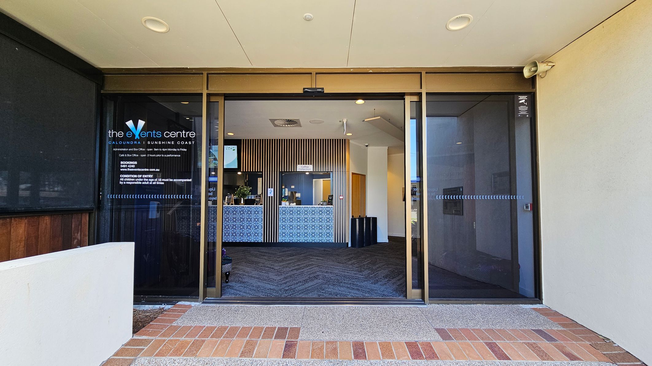 Entrance to a building with glass sliding doors. On the left, there's a sign for "the Events Centre" with details about opening hours and conditions of entry. Inside, a reception area with a patterned counter and vertical wooden slats on the wall. The floor is carpeted with a zigzag pattern. The ceiling is light-coloured with recessed lighting. The exterior floor is a mix of brick and concrete. Entrance to a building with glass sliding doors. On the left, there's a sign for "the Events Centre" with details about opening hours and conditions of entry. Inside, a reception area with a patterned counter and vertical wooden slats on the wall. The floor is carpeted with a zigzag pattern. The ceiling is light-coloured with recessed lighting. The exterior floor is a mix of brick and concrete.
