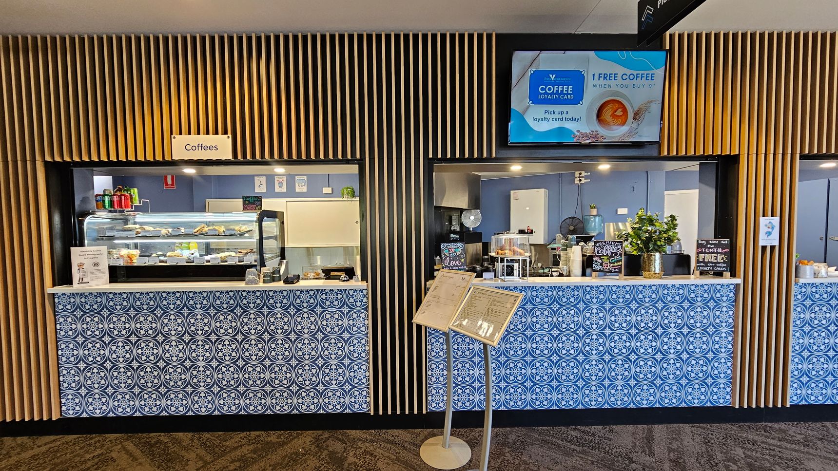 A café counter with two service windows, each adorned with blue and white patterned tiles. The left window displays a variety of snacks and drinks in a glass case, with a sign above reading "Coffees". The right window features a coffee machine and a small plant. Above, a digital screen advertises a coffee loyalty card. The background has vertical wooden slats, and a menu stand is positioned in front of the counter. The floor is carpeted with a geometric pattern. A café counter with two service windows, each adorned with blue and white patterned tiles. The left window displays a variety of snacks and drinks in a glass case, with a sign above reading "Coffees". The right window features a coffee machine and a small plant. Above, a digital screen advertises a coffee loyalty card. The background has vertical wooden slats, and a menu stand is positioned in front of the counter. The floor is carpeted with a geometric pattern.