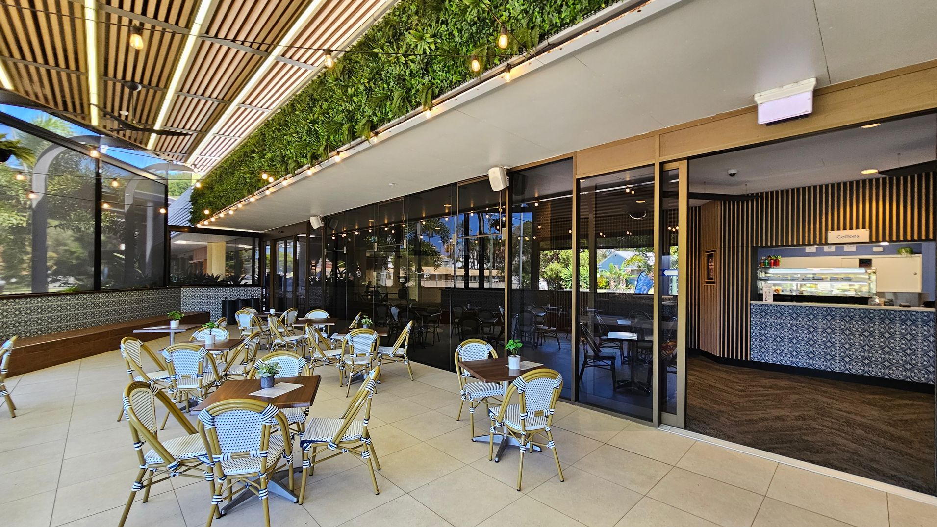 Outdoor café area with several tables and chairs on a tiled floor. On the left, a wooden bench runs along a wall with decorative tiles. The ceiling features wooden slats and string lights. A vertical garden with lush greenery is above the glass wall on the right, which separates the outdoor seating from the indoor café. Inside, a counter with patterned tiles displays a sign for coffee. Large windows reflect the surrounding greenery. Outdoor café area with several tables and chairs on a tiled floor. On the left, a wooden bench runs along a wall with decorative tiles. The ceiling features wooden slats and string lights. A vertical garden with lush greenery is above the glass wall on the right, which separates the outdoor seating from the indoor café. Inside, a counter with patterned tiles displays a sign for coffee. Large windows reflect the surrounding greenery.