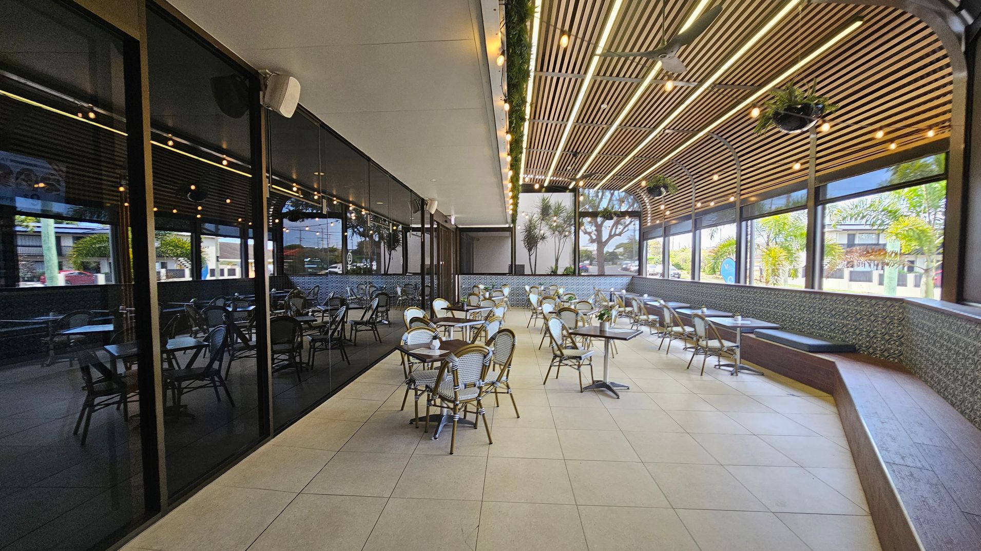 Outdoor seating area with numerous tables and chairs arranged on a tiled floor. On the left, a glass wall reflects the interior. The right side features a patterned wall with a long bench. The ceiling is wooden with hanging lights and potted plants. Large windows provide a view of trees and buildings outside. Outdoor seating area with numerous tables and chairs arranged on a tiled floor. On the left, a glass wall reflects the interior. The right side features a patterned wall with a long bench. The ceiling is wooden with hanging lights and potted plants. Large windows provide a view of trees and buildings outside.