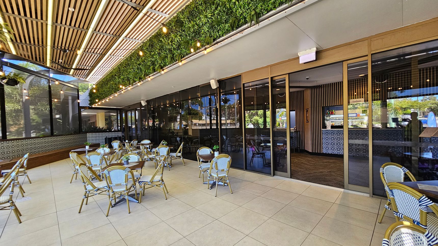 Outdoor café with wicker chairs and round tables on a tiled floor. The left side has a long bench with patterned tiles. The ceiling features wooden slats and hanging lights, with a green plant wall running along the top. Glass doors on the right lead to an indoor area with a counter and more seating. Reflections of trees and buildings are visible in the glass. Outdoor café with wicker chairs and round tables on a tiled floor. The left side has a long bench with patterned tiles. The ceiling features wooden slats and hanging lights, with a green plant wall running along the top. Glass doors on the right lead to an indoor area with a counter and more seating. Reflections of trees and buildings are visible in the glass.