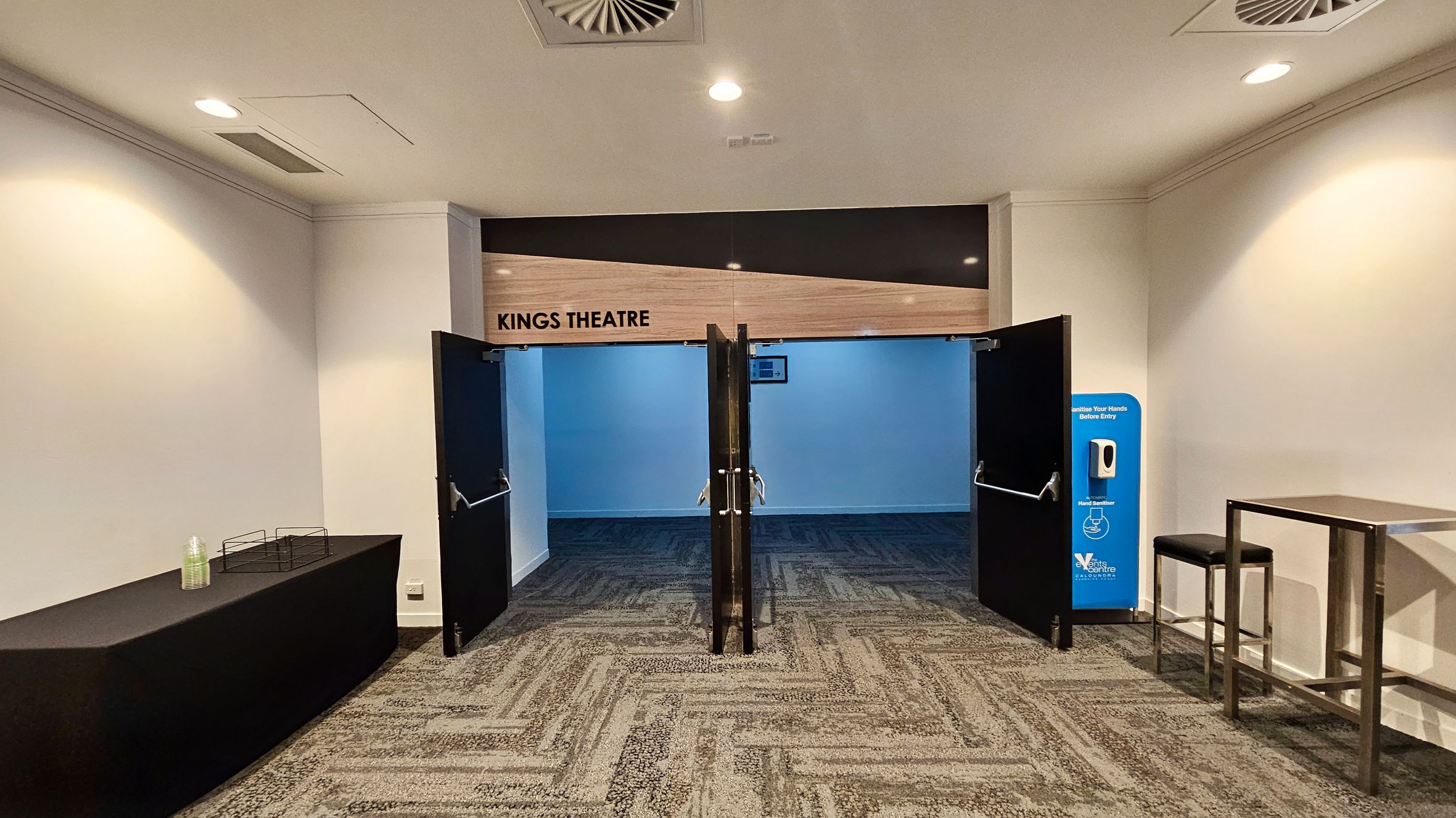 Entrance to Kings Theatre with open double doors on the left. The floor is carpeted with a geometric pattern. A small table with stools and a plant in a pot are nearby. On the right, a long bench is attached to the wall, which has several light fixtures. The ceiling is fitted with recessed lights and air vents. Further down the corridor, there are more plants and a digital display on the wall. Entrance to Kings Theatre with open double doors on the left. The floor is carpeted with a geometric pattern. A small table with stools and a plant in a pot are nearby. On the right, a long bench is attached to the wall, which has several light fixtures. The ceiling is fitted with recessed lights and air vents. Further down the corridor, there are more plants and a digital display on the wall.