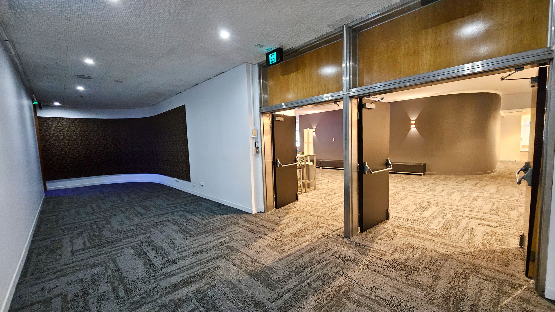 A hallway with patterned carpet flooring and a textured wall on the left. The ceiling is dotted with small lights. On the right, a set of double doors with wooden panels and metal frames is open, leading to another room with similar carpet and a curved wall. An illuminated exit sign is above the doors. The room beyond has a wall light fixture and some furniture, including a bench and a table. A hallway with patterned carpet flooring and a textured wall on the left. The ceiling is dotted with small lights. On the right, a set of double doors with wooden panels and metal frames is open, leading to another room with similar carpet and a curved wall. An illuminated exit sign is above the doors. The room beyond has a wall light fixture and some furniture, including a bench and a table.