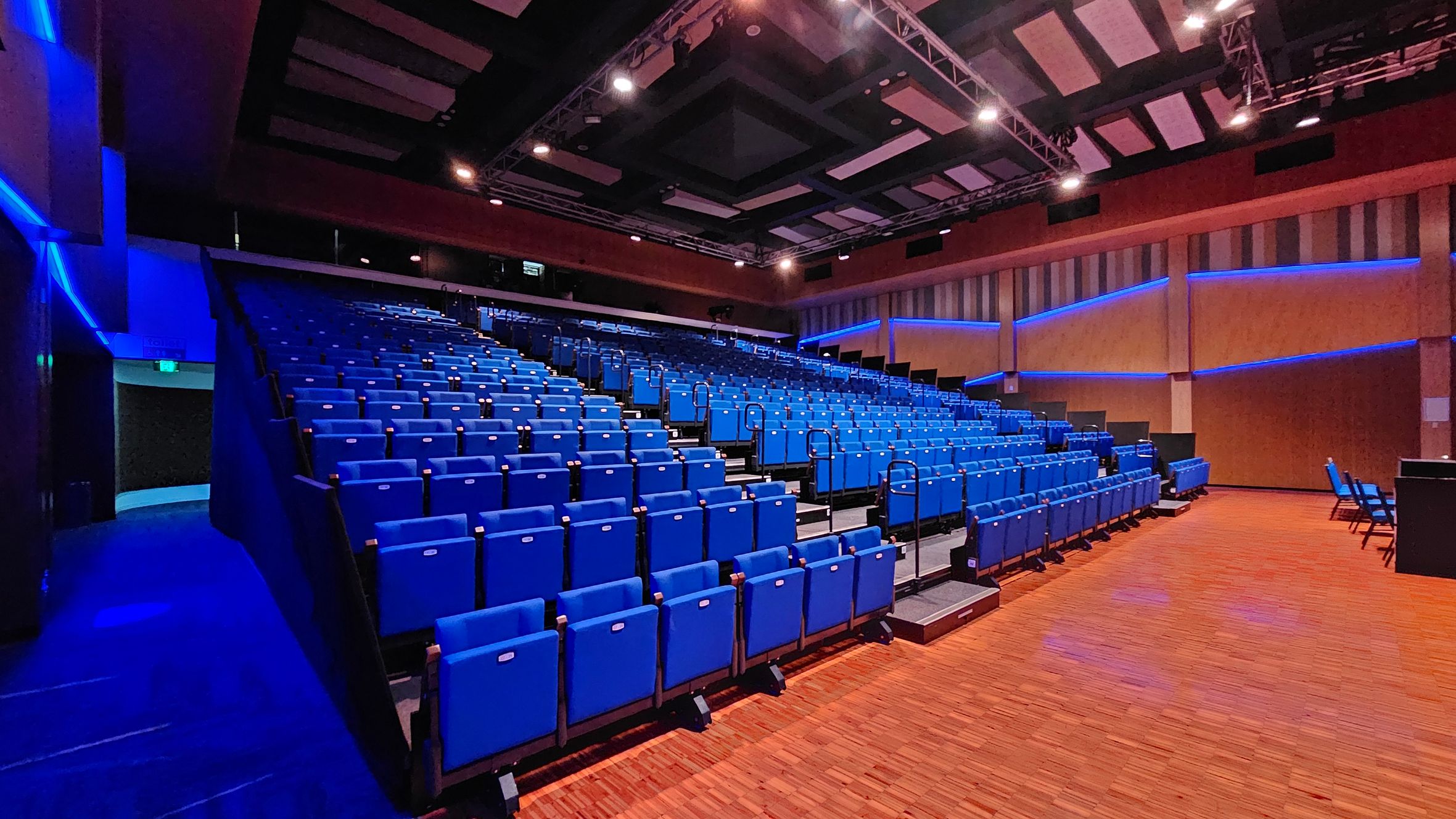 An auditorium with rows of blue seats arranged in tiers on the left and centre. The floor is wooden with a warm tone. The walls are lined with vertical panels and illuminated by blue LED lights. The ceiling features a grid of acoustic panels and spotlights. On the right, there is a small area with additional seating and a table. An auditorium with rows of blue seats arranged in tiers on the left and centre. The floor is wooden with a warm tone. The walls are lined with vertical panels and illuminated by blue LED lights. The ceiling features a grid of acoustic panels and spotlights. On the right, there is a small area with additional seating and a table.