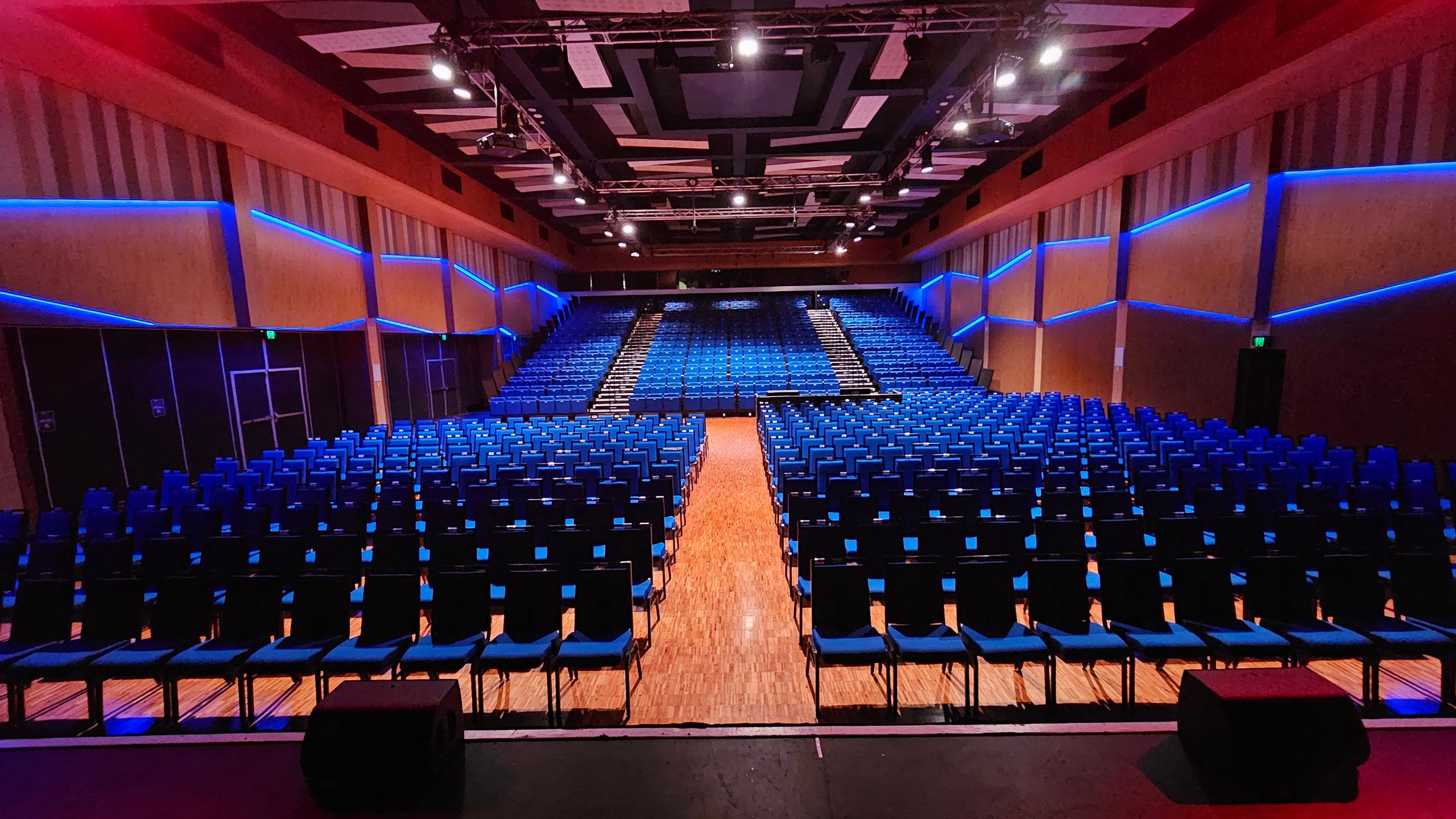 A large auditorium with rows of blue chairs arranged in a semi-circular pattern. The walls are lined with blue LED lights, creating a modern look. The ceiling features a geometric design with spotlights. The floor is wooden, and there are aisles dividing the seating sections. The view is from the stage, looking towards the back of the auditorium. A large auditorium with rows of blue chairs arranged in a semi-circular pattern. The walls are lined with blue LED lights, creating a modern look. The ceiling features a geometric design with spotlights. The floor is wooden, and there are aisles dividing the seating sections. The view is from the stage, looking towards the back of the auditorium.