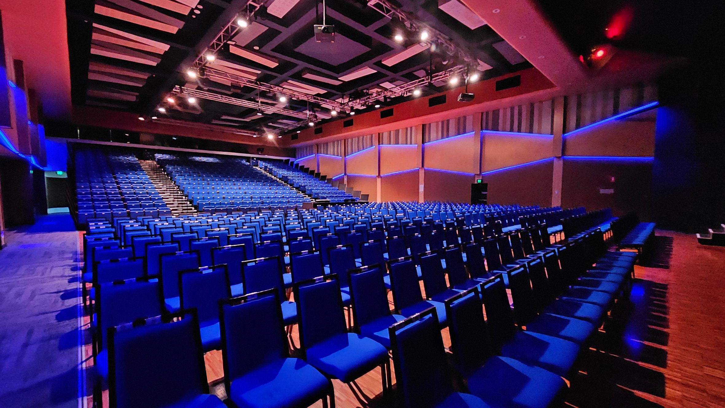 Rows of blue chairs fill a large auditorium, with a central aisle leading up to a stage. The walls are adorned with geometric patterns and blue lighting accents. The ceiling features a complex grid of lights and panels. The floor is wooden, and the space is well-lit, creating a vibrant atmosphere. Rows of blue chairs fill a large auditorium, with a central aisle leading up to a stage. The walls are adorned with geometric patterns and blue lighting accents. The ceiling features a complex grid of lights and panels. The floor is wooden, and the space is well-lit, creating a vibrant atmosphere.