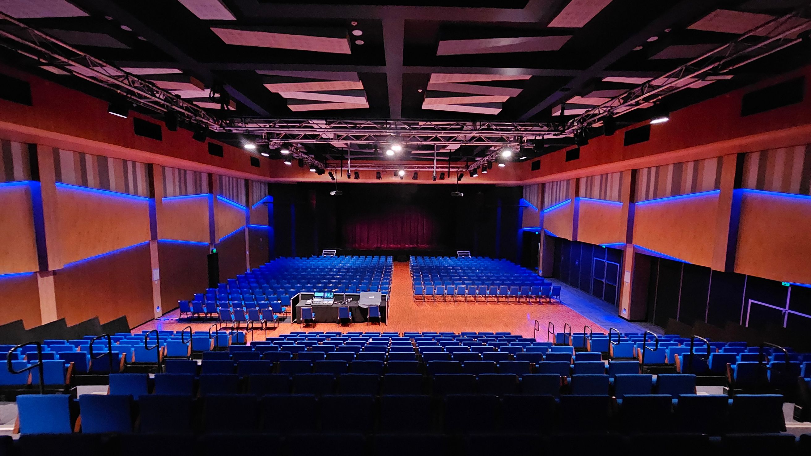 A large auditorium with rows of blue seats facing a stage at the back. The stage is framed by dark curtains. In the centre, there is a control desk with equipment and chairs. The walls are lined with vertical panels and illuminated by blue lights. The ceiling features a grid of beams and spotlights. The floor is wooden, and there are handrails along the aisles. A large auditorium with rows of blue seats facing a stage at the back. The stage is framed by dark curtains. In the centre, there is a control desk with equipment and chairs. The walls are lined with vertical panels and illuminated by blue lights. The ceiling features a grid of beams and spotlights. The floor is wooden, and there are handrails along the aisles.