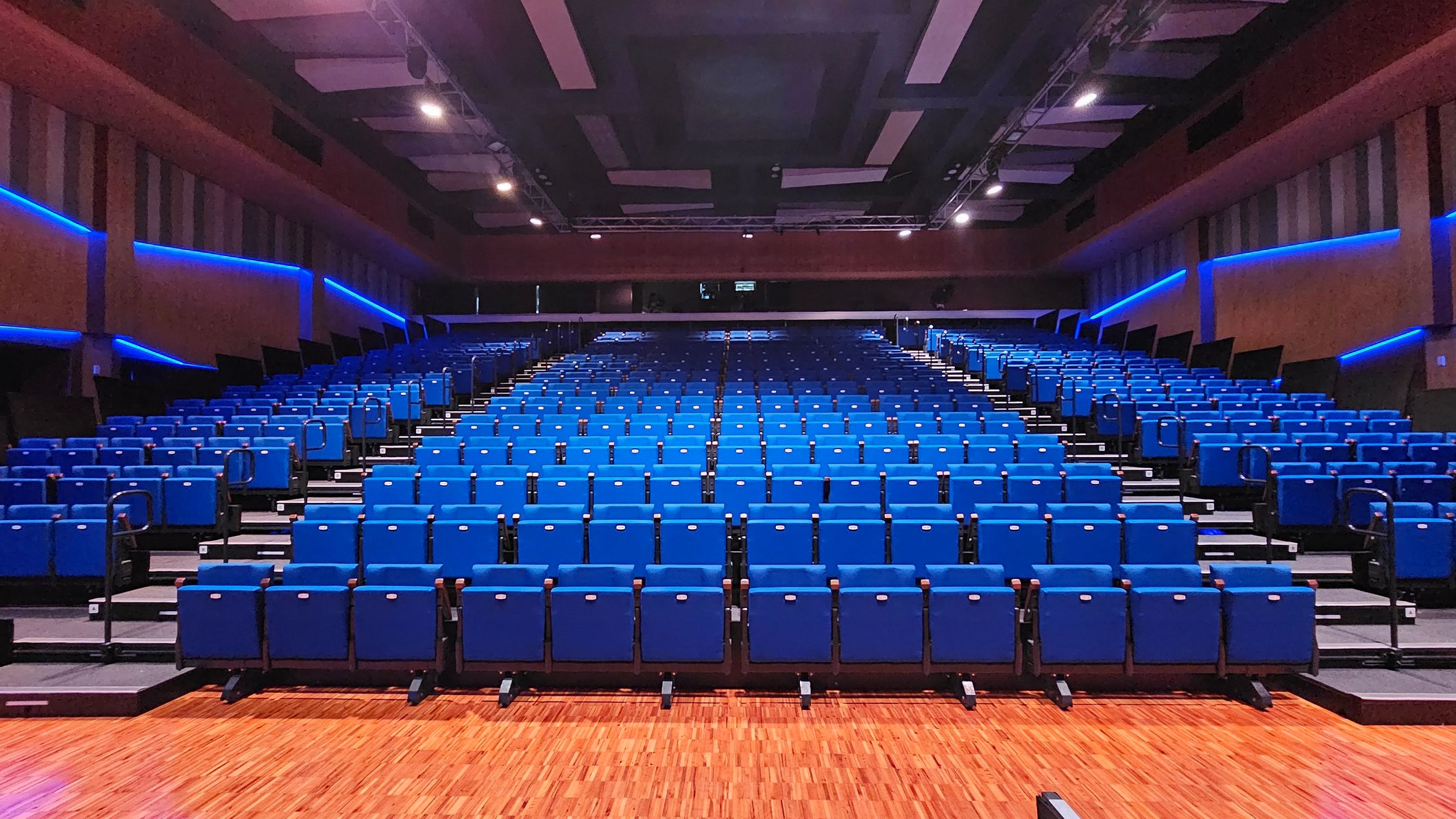 Rows of blue theatre seats are arranged in a large auditorium. The seats are on a sloped floor, leading up to the back of the room. The walls are lined with blue LED lights, creating a modern atmosphere. The ceiling is equipped with rectangular panels and spotlights. The floor is wooden, adding warmth to the space. Rows of blue theatre seats are arranged in a large auditorium. The seats are on a sloped floor, leading up to the back of the room. The walls are lined with blue LED lights, creating a modern atmosphere. The ceiling is equipped with rectangular panels and spotlights. The floor is wooden, adding warmth to the space.