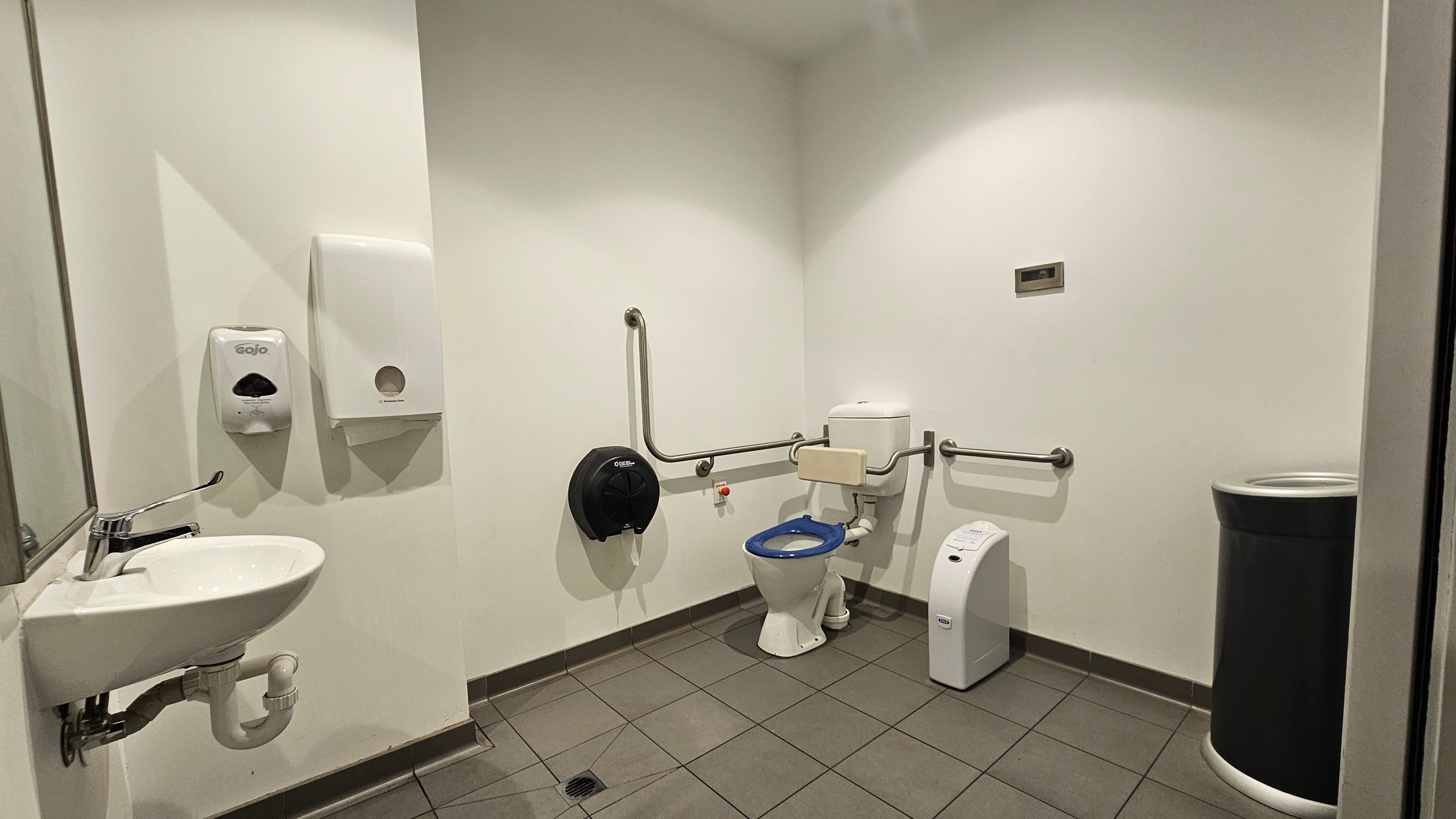 Accessible bathroom with a white sink and a mirror on the left wall. Above the sink are a soap dispenser and a paper towel dispenser. In the centre, a toilet with a blue seat is equipped with support rails on both sides. A black toilet paper dispenser is mounted on the wall beside it. On the right, a sanitary bin is placed against the wall. The floor is tiled in a light brown colour, and the walls are painted white. Accessible bathroom with a white sink and a mirror on the left wall. Above the sink are a soap dispenser and a paper towel dispenser. In the centre, a toilet with a blue seat is equipped with support rails on both sides. A black toilet paper dispenser is mounted on the wall beside it. On the right, a sanitary bin is placed against the wall. The floor is tiled in a light brown colour, and the walls are painted white.