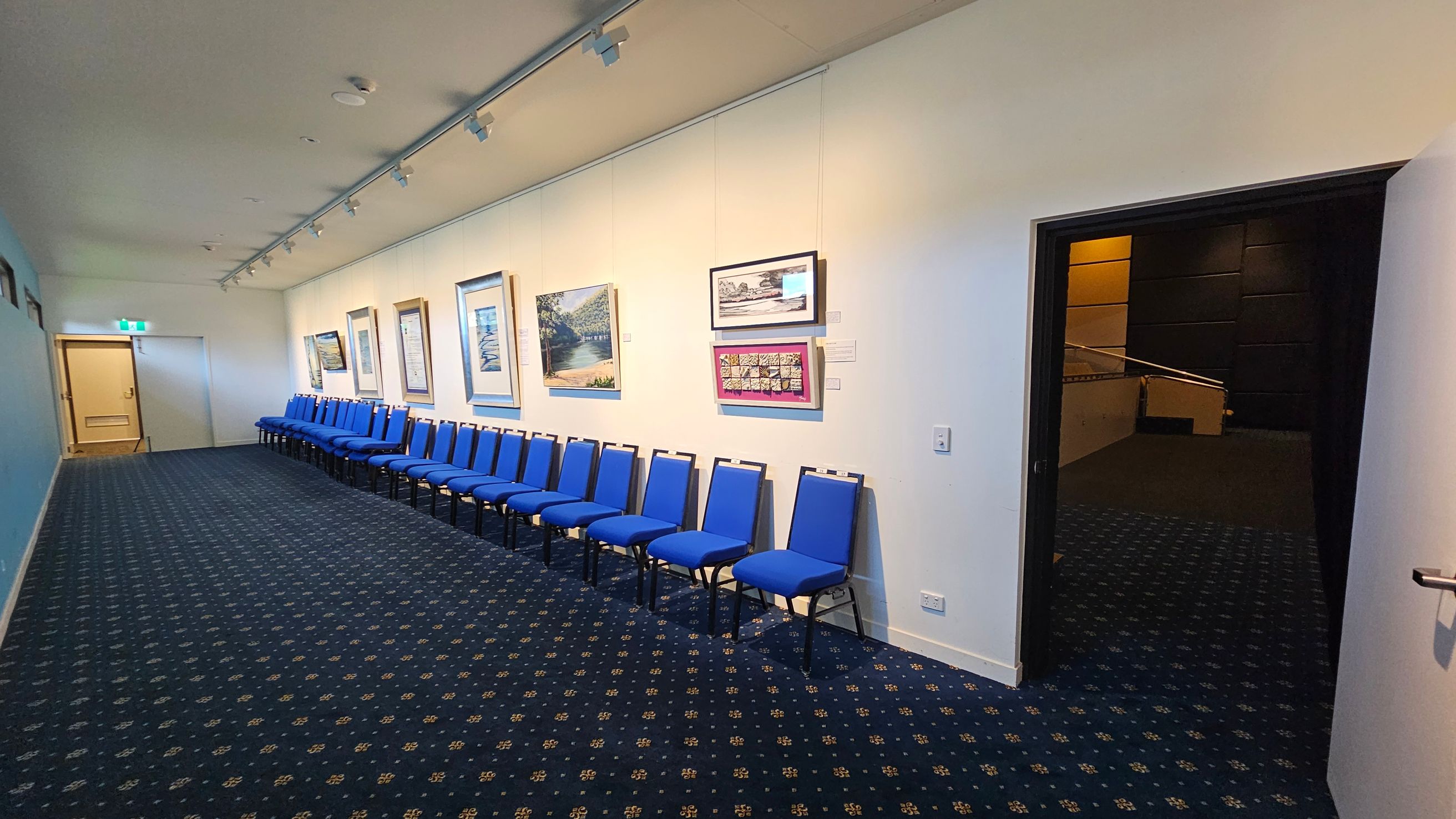 A hallway with a sign reading "Playhouse Theatre" above a set of carpeted stairs leading upwards. On the left wall, there is a sign with arrows indicating directions to the toilets and accessible facilities. On the right, a tall table with a black stool and a colourful cushion on the floor. The floor is carpeted with a patterned design, and the walls are painted in light colours. A hallway with a sign reading "Playhouse Theatre" above a set of carpeted stairs leading upwards. On the left wall, there is a sign with arrows indicating directions to the toilets and accessible facilities. On the right, a tall table with a black stool and a colourful cushion on the floor. The floor is carpeted with a patterned design, and the walls are painted in light colours.