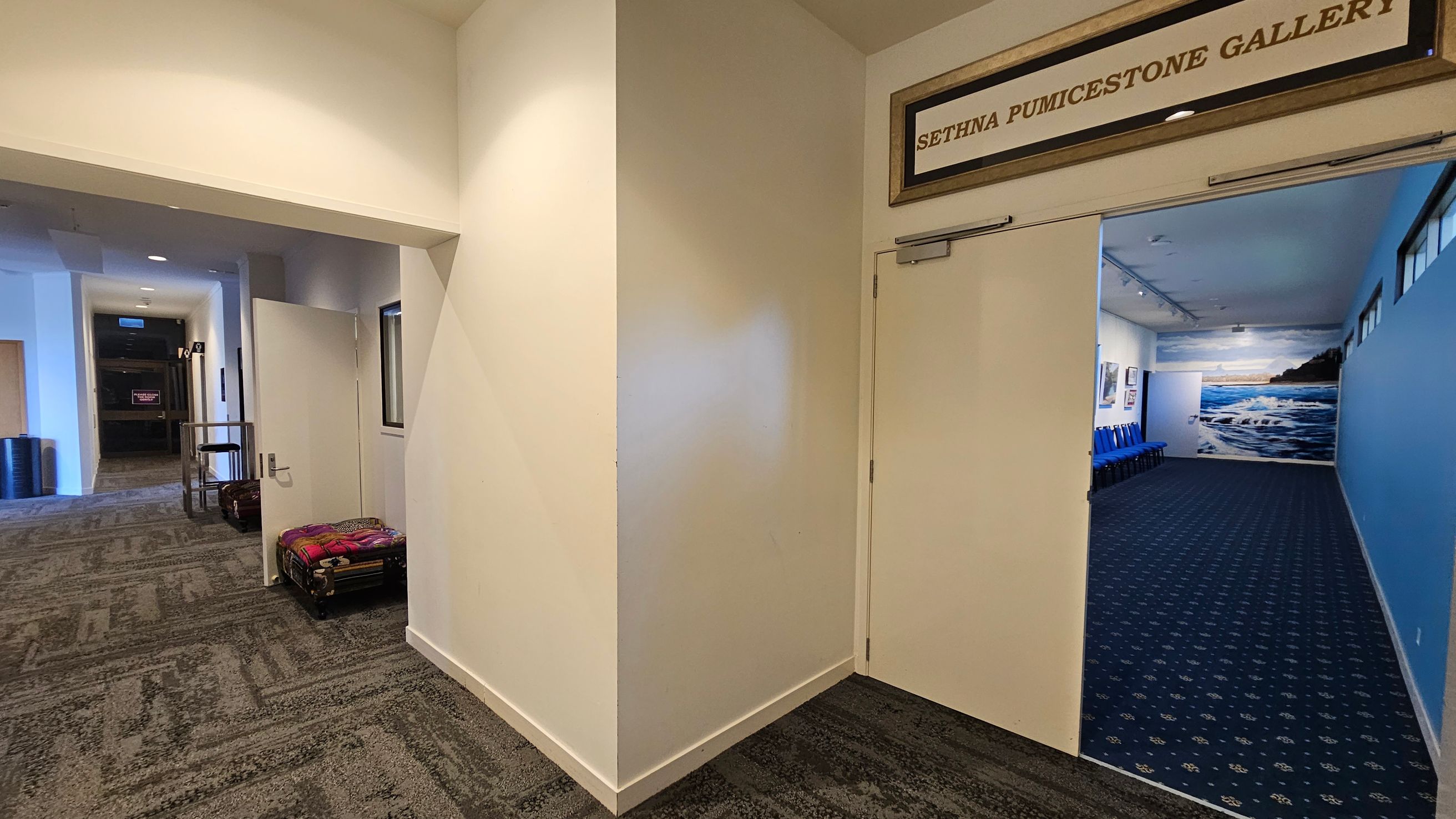 A hallway with a patterned carpet leads to an open door on the right, marked "Sethna Pumicestone Gallery." Inside, the gallery has blue walls and a mural of a seascape. Blue chairs are lined up against the left wall. On the left side of the hallway, there is a partially open door and a trolley with luggage. The ceiling has recessed lighting, and the walls are painted in a light colour. A hallway with a patterned carpet leads to an open door on the right, marked "Sethna Pumicestone Gallery." Inside, the gallery has blue walls and a mural of a seascape. Blue chairs are lined up against the left wall. On the left side of the hallway, there is a partially open door and a trolley with luggage. The ceiling has recessed lighting, and the walls are painted in a light colour.