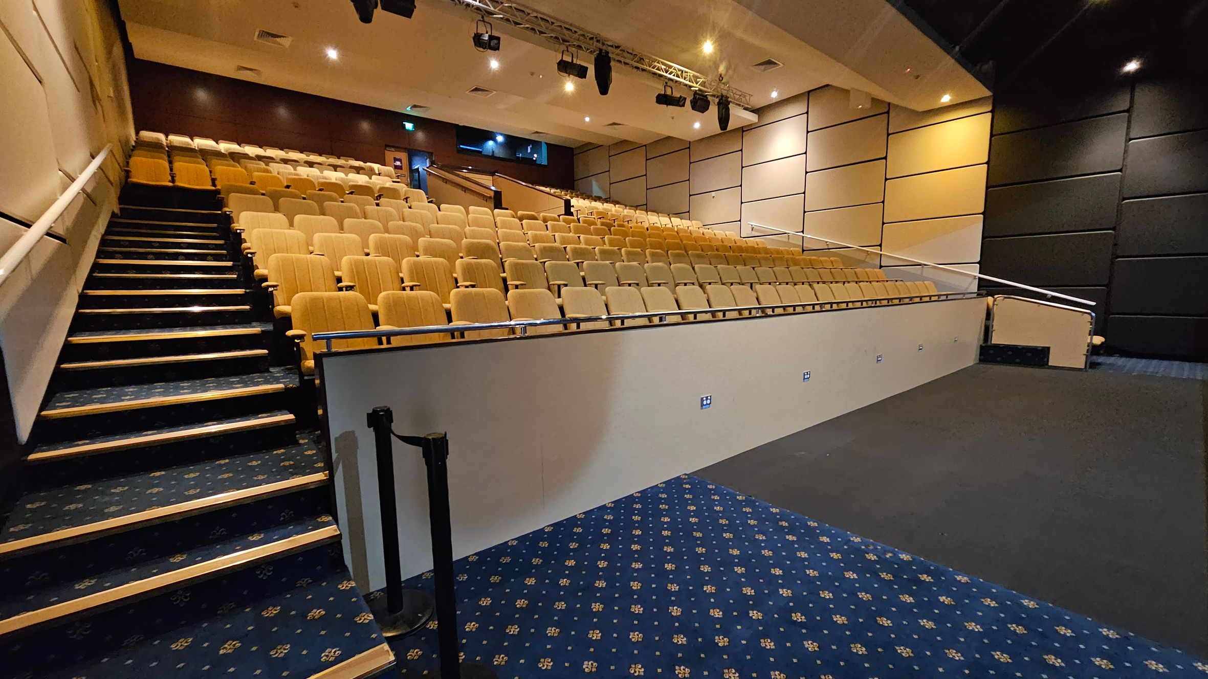A lecture hall with tiered seating on the left, featuring rows of beige chairs. A staircase with a dark blue patterned carpet runs alongside the seats. The right side has a wall with beige and black acoustic panels. The floor in the foreground is a mix of dark blue patterned carpet and plain dark flooring. The ceiling is equipped with spotlights and a lighting rig. A lecture hall with tiered seating on the left, featuring rows of beige chairs. A staircase with a dark blue patterned carpet runs alongside the seats. The right side has a wall with beige and black acoustic panels. The floor in the foreground is a mix of dark blue patterned carpet and plain dark flooring. The ceiling is equipped with spotlights and a lighting rig.
