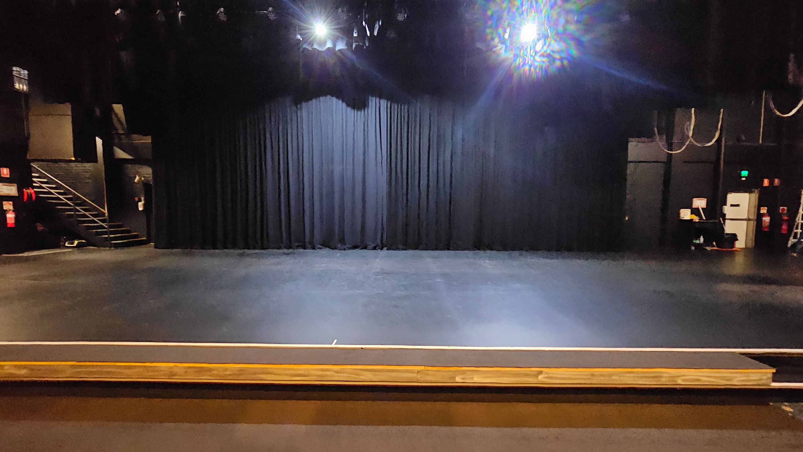 A theatre stage with a black curtain backdrop. The floor is dark and smooth, extending to the front of the stage. On the left, there is a staircase leading up to a higher level. On the right, there are some technical equipment and a ladder. Overhead, stage lights are visible, casting a bright glow. The ceiling is not visible. A theatre stage with a black curtain backdrop. The floor is dark and smooth, extending to the front of the stage. On the left, there is a staircase leading up to a higher level. On the right, there are some technical equipment and a ladder. Overhead, stage lights are visible, casting a bright glow. The ceiling is not visible.
