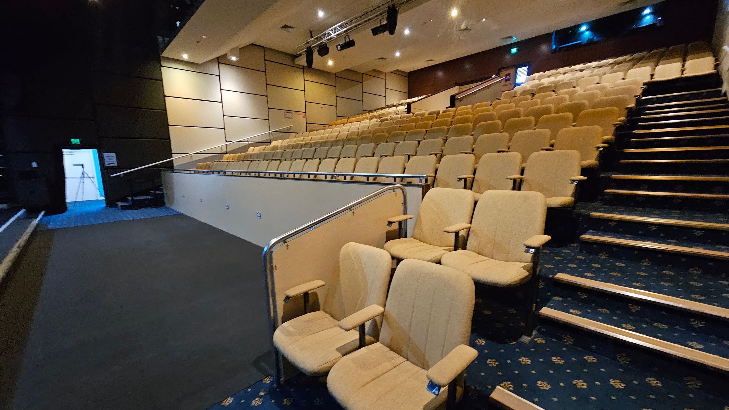 A lecture hall with tiered seating on the right, featuring beige upholstered chairs. The floor is carpeted in a dark blue pattern. On the left, there is a walkway with a metal railing and a door with a blue light visible through it. The walls are panelled in a light colour, and the ceiling has recessed lighting and mounted speakers. A lecture hall with tiered seating on the right, featuring beige upholstered chairs. The floor is carpeted in a dark blue pattern. On the left, there is a walkway with a metal railing and a door with a blue light visible through it. The walls are panelled in a light colour, and the ceiling has recessed lighting and mounted speakers.