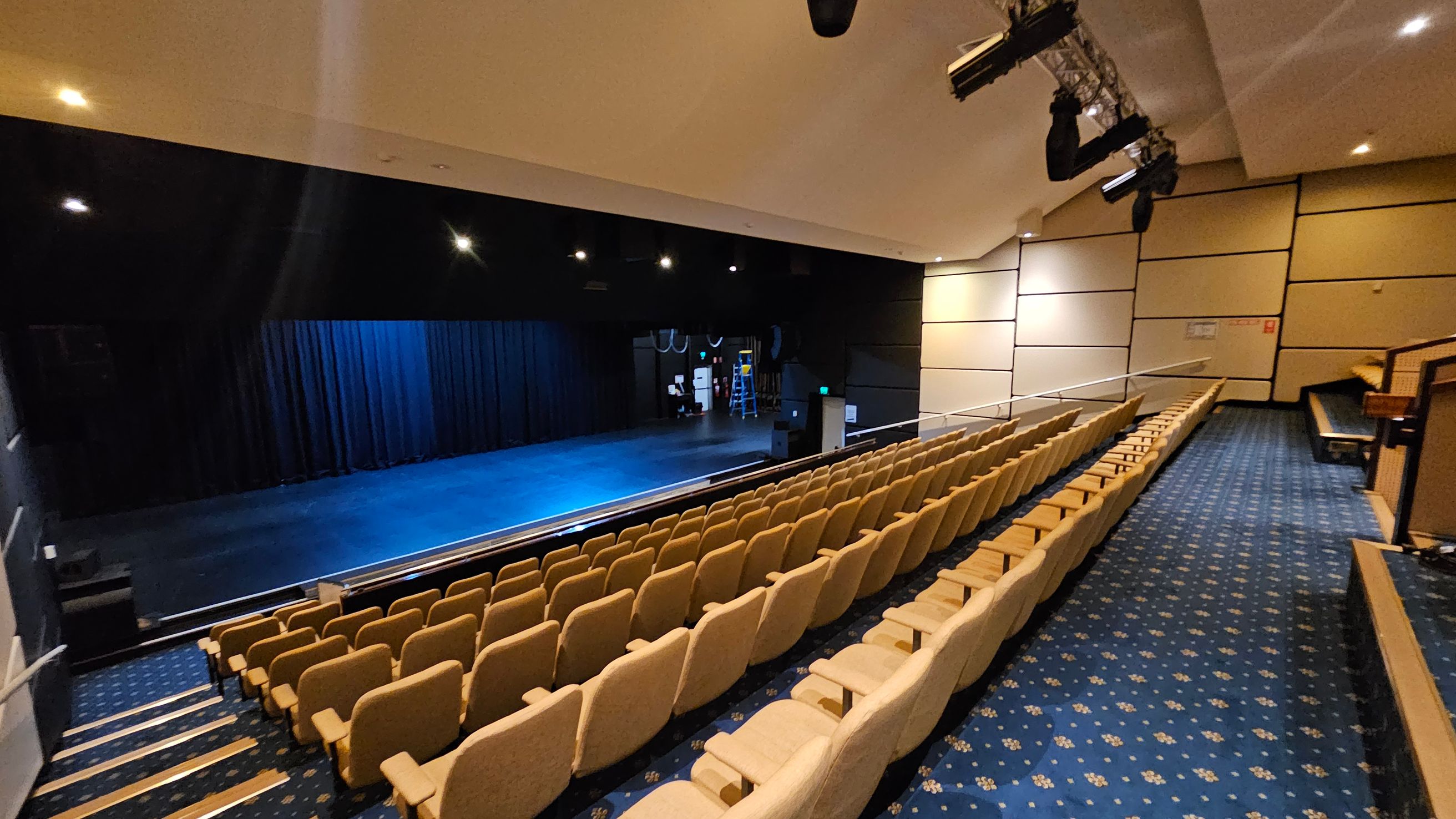 A theatre interior with rows of beige seats on the right, arranged in a tiered fashion. The floor is covered with a blue carpet featuring a floral pattern. On the left is a stage with a dark backdrop and blue lighting. Above the stage, several stage lights are mounted. The walls are lined with beige panels, and the ceiling is equipped with recessed lighting. A theatre interior with rows of beige seats on the right, arranged in a tiered fashion. The floor is covered with a blue carpet featuring a floral pattern. On the left is a stage with a dark backdrop and blue lighting. Above the stage, several stage lights are mounted. The walls are lined with beige panels, and the ceiling is equipped with recessed lighting.
