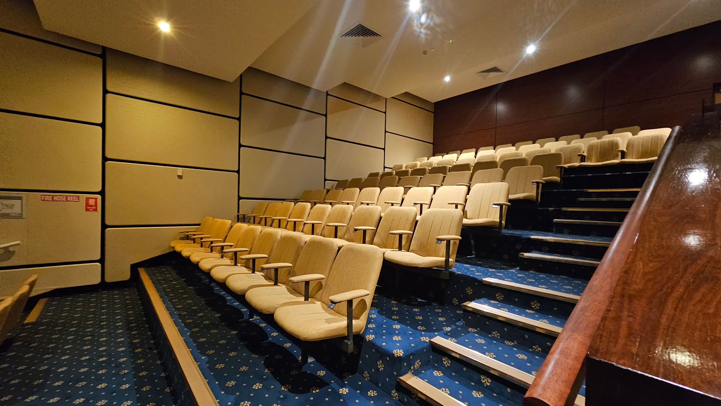 A small auditorium with tiered seating. Beige upholstered chairs are arranged in rows on a blue carpet with a floral pattern. The left wall is covered with large, rectangular acoustic panels, and a fire hose reel sign is visible. The right side features a wooden handrail along the stairs. The ceiling is angled with recessed lighting. A small auditorium with tiered seating. Beige upholstered chairs are arranged in rows on a blue carpet with a floral pattern. The left wall is covered with large, rectangular acoustic panels, and a fire hose reel sign is visible. The right side features a wooden handrail along the stairs. The ceiling is angled with recessed lighting.