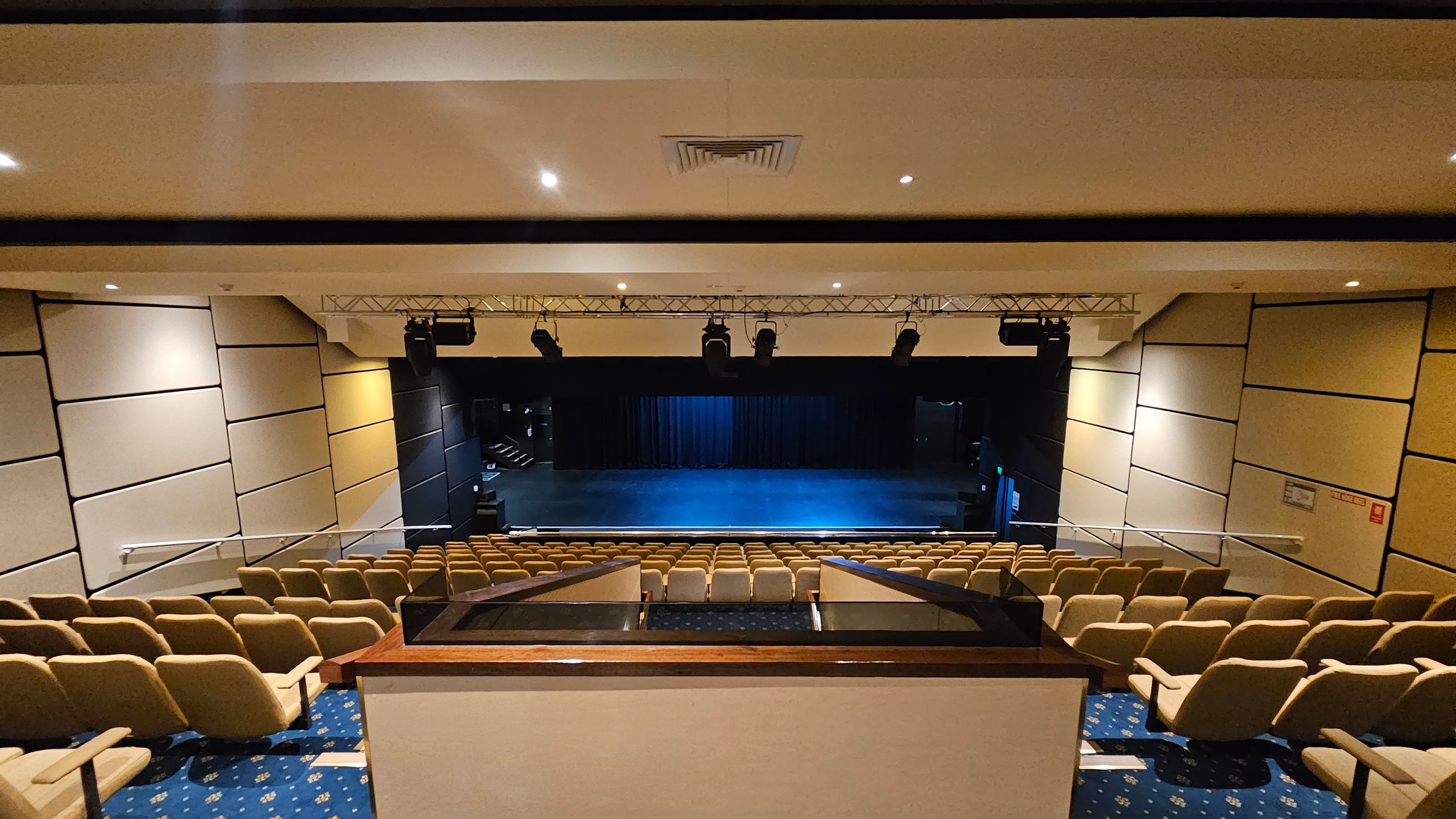 A theatre interior with rows of beige seats facing a stage. The stage is empty with a blue-lit floor and a black curtain backdrop. Overhead, there are stage lights mounted on a metal truss. The walls are lined with beige acoustic panels. The floor is carpeted in blue with a pattern, and the ceiling is white with recessed lighting and a central air vent. A theatre interior with rows of beige seats facing a stage. The stage is empty with a blue-lit floor and a black curtain backdrop. Overhead, there are stage lights mounted on a metal truss. The walls are lined with beige acoustic panels. The floor is carpeted in blue with a pattern, and the ceiling is white with recessed lighting and a central air vent.