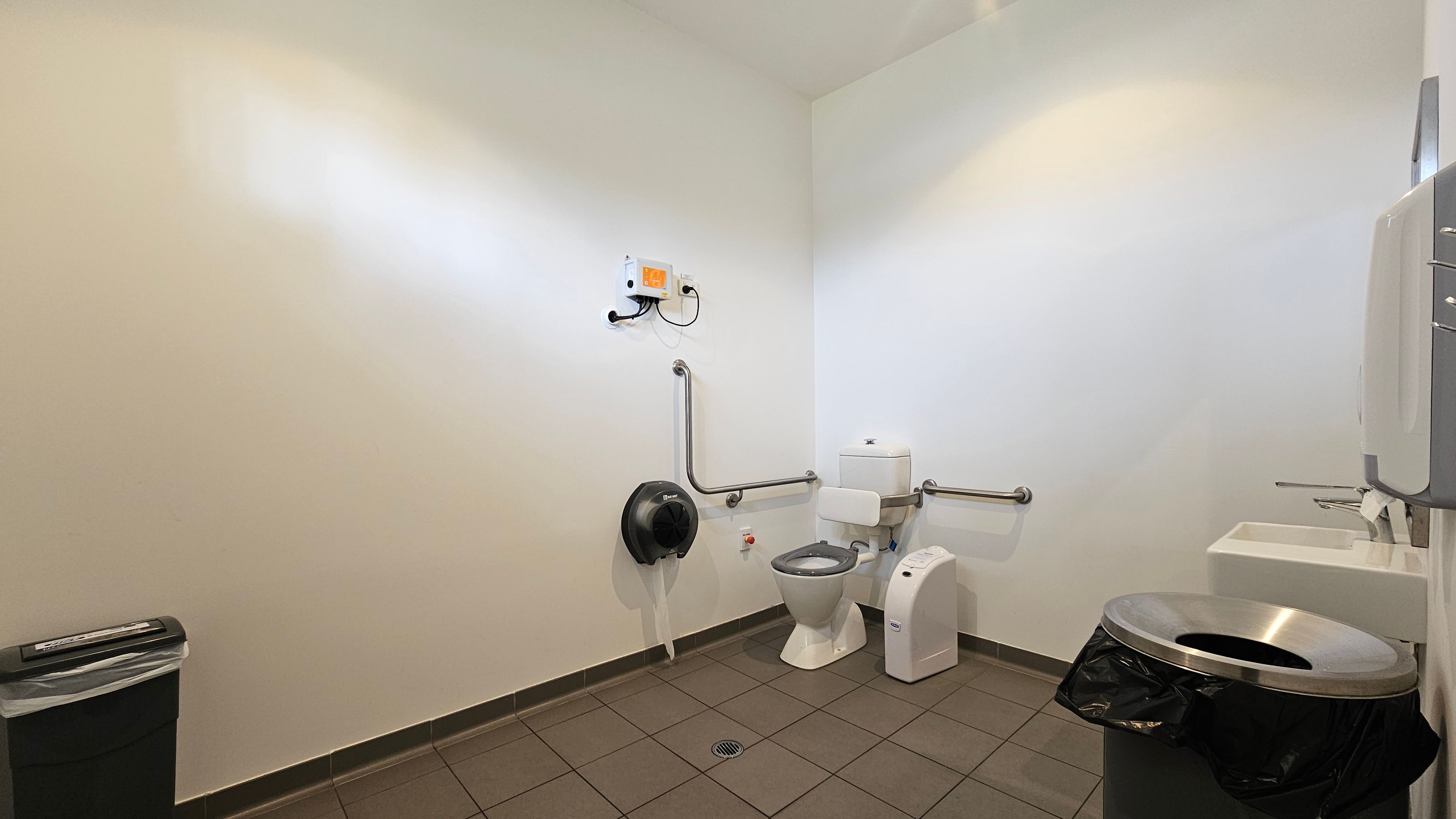 A public restroom with a white tiled floor and white walls. On the right, there's a sink with a soap dispenser above it. Next to the sink is a toilet with support rails on both sides. Above the toilet, a small white box is mounted on the wall. To the left of the toilet, a large toilet paper dispenser is attached to the wall. In the foreground on the left, a black bin with a silver lid is visible. A public restroom with a white tiled floor and white walls. On the right, there's a sink with a soap dispenser above it. Next to the sink is a toilet with support rails on both sides. Above the toilet, a small white box is mounted on the wall. To the left of the toilet, a large toilet paper dispenser is attached to the wall. In the foreground on the left, a black bin with a silver lid is visible.