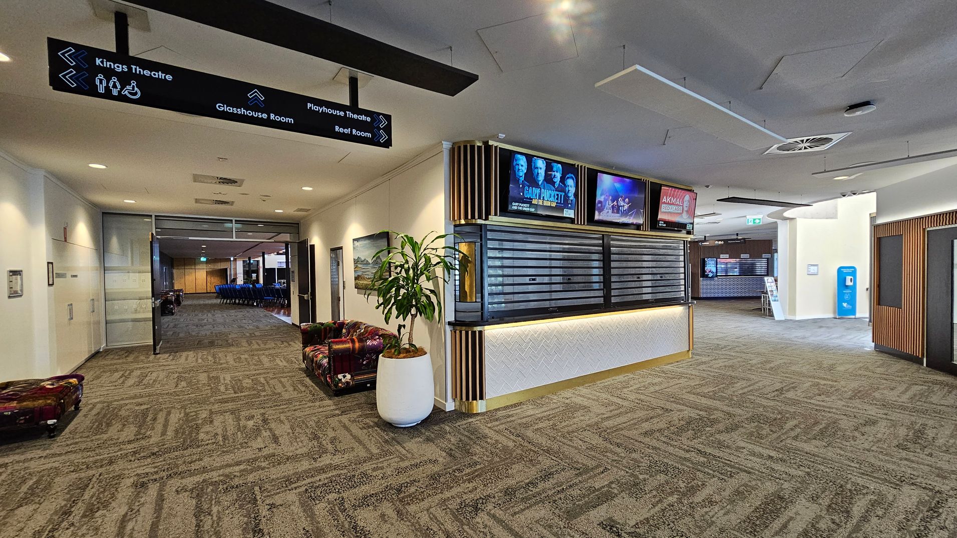 A spacious lobby area with a patterned carpet floor. On the left, there is a hallway leading to a room with blue chairs, and a sign above pointing to various locations like Kings Theatre and Glasshouse Room. A potted plant sits near a colourful cushioned bench. In the centre, a counter with digital screens above displays event information. On the right, a blue vending machine is visible near another hallway. The ceiling has recessed lighting and air vents. A spacious lobby area with a patterned carpet floor. On the left, there is a hallway leading to a room with blue chairs, and a sign above pointing to various locations like Kings Theatre and Glasshouse Room. A potted plant sits near a colourful cushioned bench. In the centre, a counter with digital screens above displays event information. On the right, a blue vending machine is visible near another hallway. The ceiling has recessed lighting and air vents.