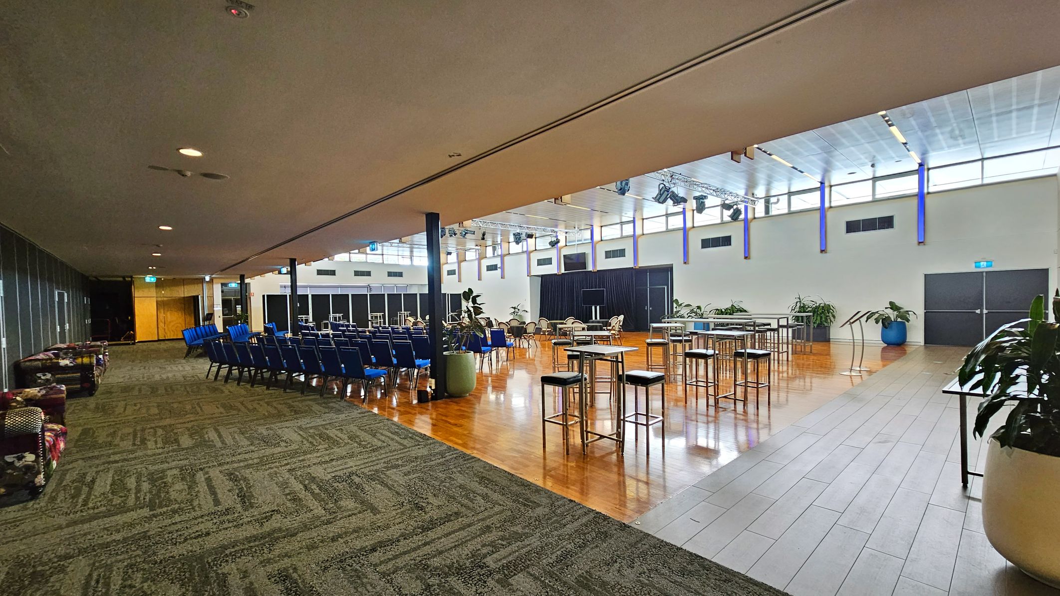 A spacious indoor venue with a mix of carpeted and wooden flooring. On the left, there are rows of blue chairs facing a stage area. The right side features high tables with stools and potted plants. The ceiling is high with large windows allowing natural light. The walls are white with blue vertical accents, and there are doors at the far end. The ceiling has lighting fixtures and speakers. A spacious indoor venue with a mix of carpeted and wooden flooring. On the left, there are rows of blue chairs facing a stage area. The right side features high tables with stools and potted plants. The ceiling is high with large windows allowing natural light. The walls are white with blue vertical accents, and there are doors at the far end. The ceiling has lighting fixtures and speakers.