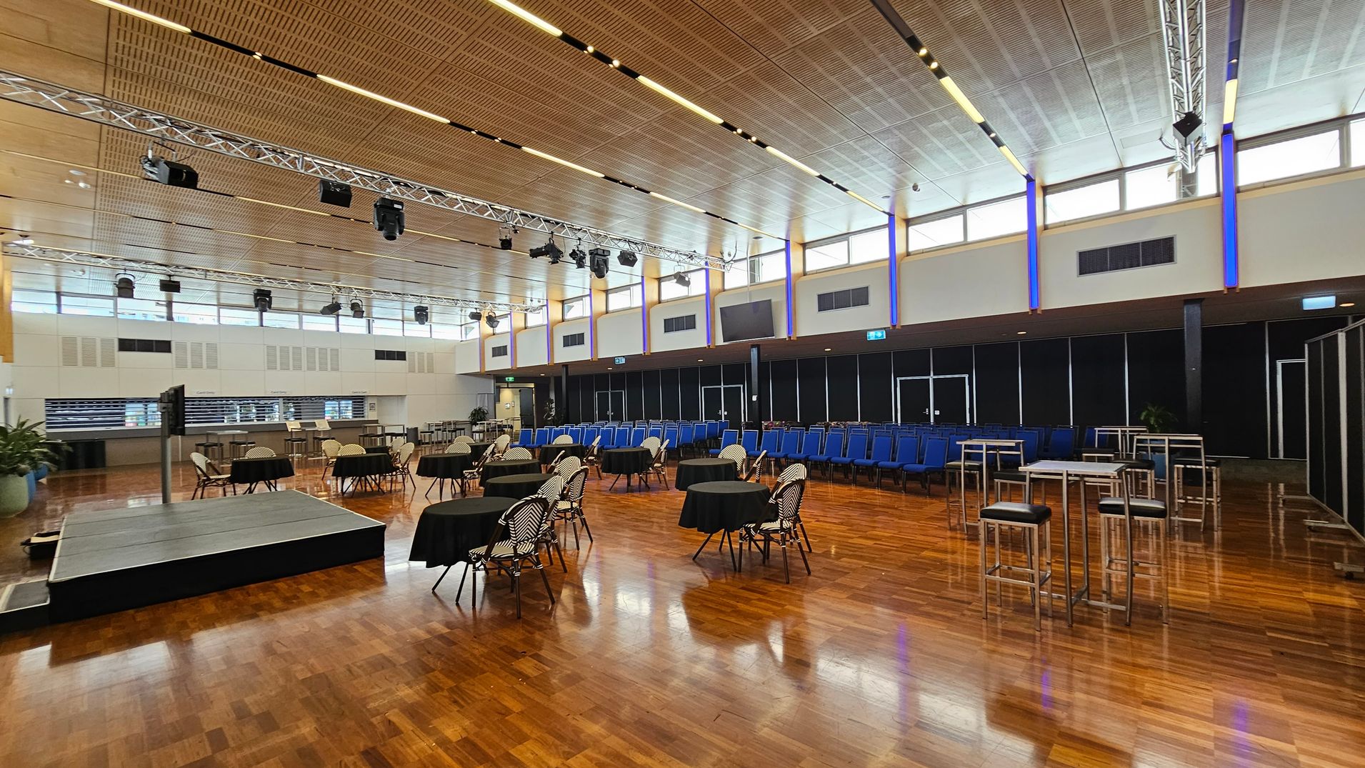 A spacious indoor venue with rows of blue chairs arranged in the centre on a wooden floor. To the left, there are tall tables and chairs, along with potted plants. The right side features a carpeted area with additional seating and tables. The ceiling is high with visible lighting fixtures, and large windows allow natural light to enter. In the background, there are more plants and a dark curtain. The far right shows an open corridor leading to another area. A spacious indoor venue with rows of blue chairs arranged in the centre on a wooden floor. To the left, there are tall tables and chairs, along with potted plants. The right side features a carpeted area with additional seating and tables. The ceiling is high with visible lighting fixtures, and large windows allow natural light to enter. In the background, there are more plants and a dark curtain. The far right shows an open corridor leading to another area.