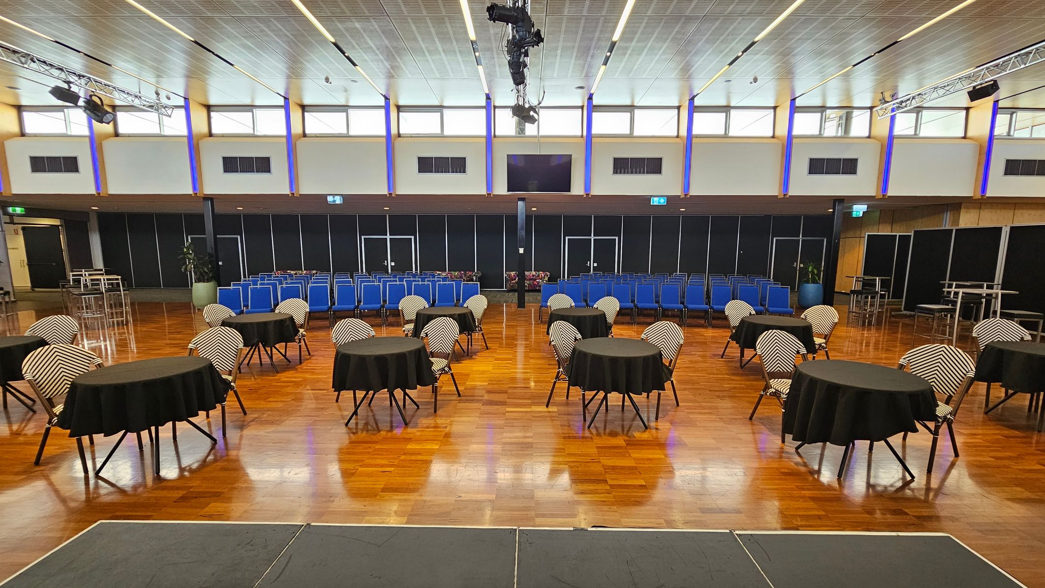 A spacious hall with a wooden floor and a high, panelled ceiling. On the left, there is a small stage and several round tables with black tablecloths and chairs. The right side features rows of blue chairs and a few tall tables with stools. The back wall has large windows and black panels, with lighting fixtures and speakers suspended from the ceiling. A spacious hall with a wooden floor and a high, panelled ceiling. On the left, there is a small stage and several round tables with black tablecloths and chairs. The right side features rows of blue chairs and a few tall tables with stools. The back wall has large windows and black panels, with lighting fixtures and speakers suspended from the ceiling.