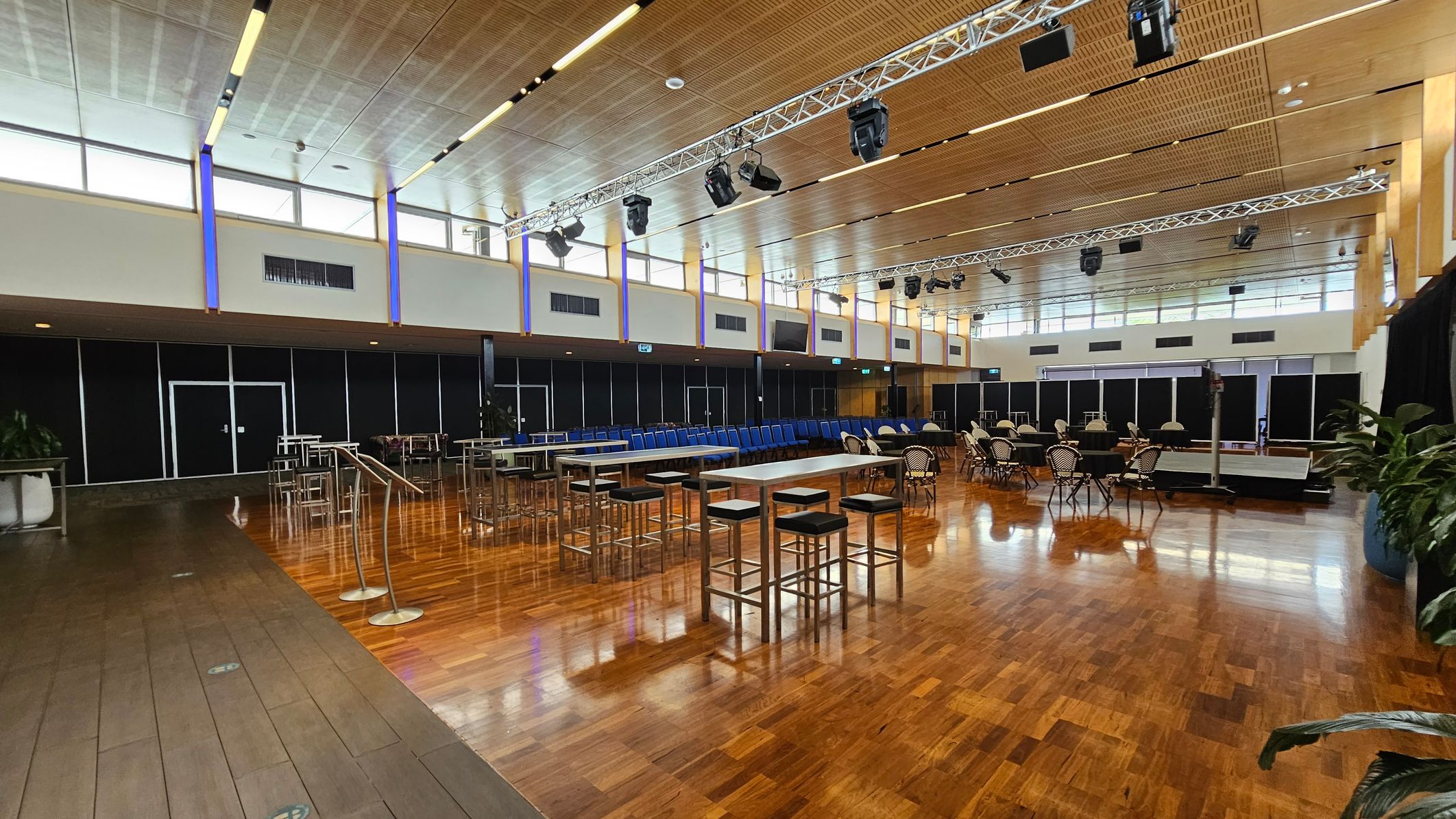 A spacious hall with a polished wooden floor. In the foreground, several round tables are covered with black tablecloths, surrounded by chairs with black and white striped covers. In the background, rows of blue chairs are neatly arranged. The ceiling is high with a grid pattern and features lighting fixtures. The walls have large windows near the ceiling, allowing natural light to enter. On the sides, there are tall plants in pots and some high tables with stools. A spacious hall with a polished wooden floor. In the foreground, several round tables are covered with black tablecloths, surrounded by chairs with black and white striped covers. In the background, rows of blue chairs are neatly arranged. The ceiling is high with a grid pattern and features lighting fixtures. The walls have large windows near the ceiling, allowing natural light to enter. On the sides, there are tall plants in pots and some high tables with stools.