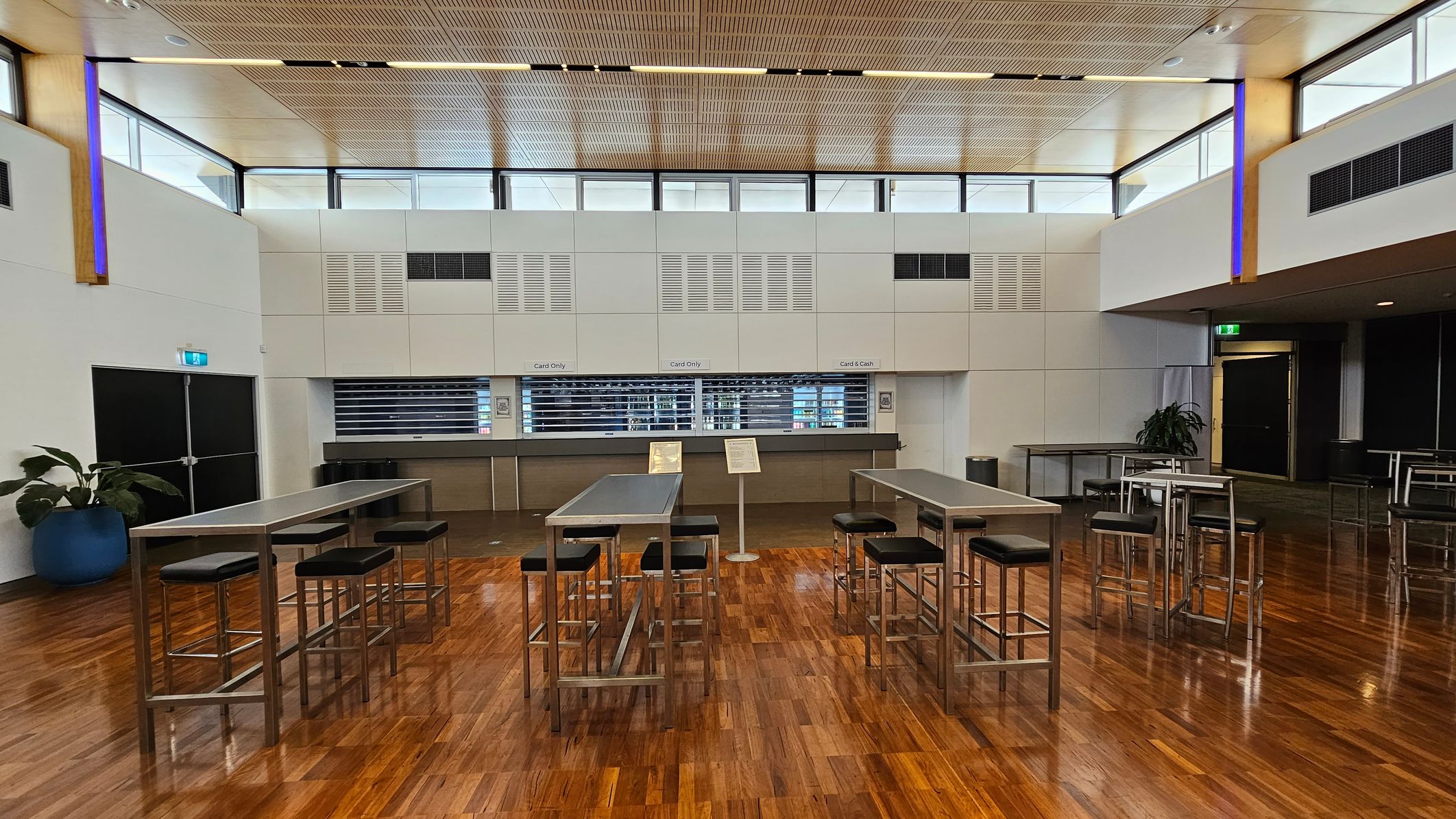 A spacious indoor venue with a polished wooden floor. On the left, there are high tables with stools and a few potted plants. In the centre, rows of blue chairs are arranged in front of a stage area. On the right, round tables with chairs are scattered. The ceiling is high with wooden panels and equipped with lighting and speakers. Large windows near the ceiling allow natural light to enter. A spacious indoor venue with a polished wooden floor. On the left, there are high tables with stools and a few potted plants. In the centre, rows of blue chairs are arranged in front of a stage area. On the right, round tables with chairs are scattered. The ceiling is high with wooden panels and equipped with lighting and speakers. Large windows near the ceiling allow natural light to enter.