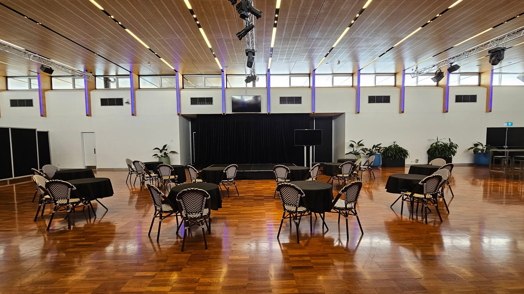 A spacious room with a polished wooden floor and a high ceiling featuring recessed lighting. In the centre, there are several round tables covered with black tablecloths, each surrounded by chairs with a woven pattern. At the back, a small stage is set against a black curtain. Potted plants are placed along the walls, and a large screen is visible to the right. The walls have high windows, allowing natural light to enter. A spacious room with a polished wooden floor and a high ceiling featuring recessed lighting. In the centre, there are several round tables covered with black tablecloths, each surrounded by chairs with a woven pattern. At the back, a small stage is set against a black curtain. Potted plants are placed along the walls, and a large screen is visible to the right. The walls have high windows, allowing natural light to enter.