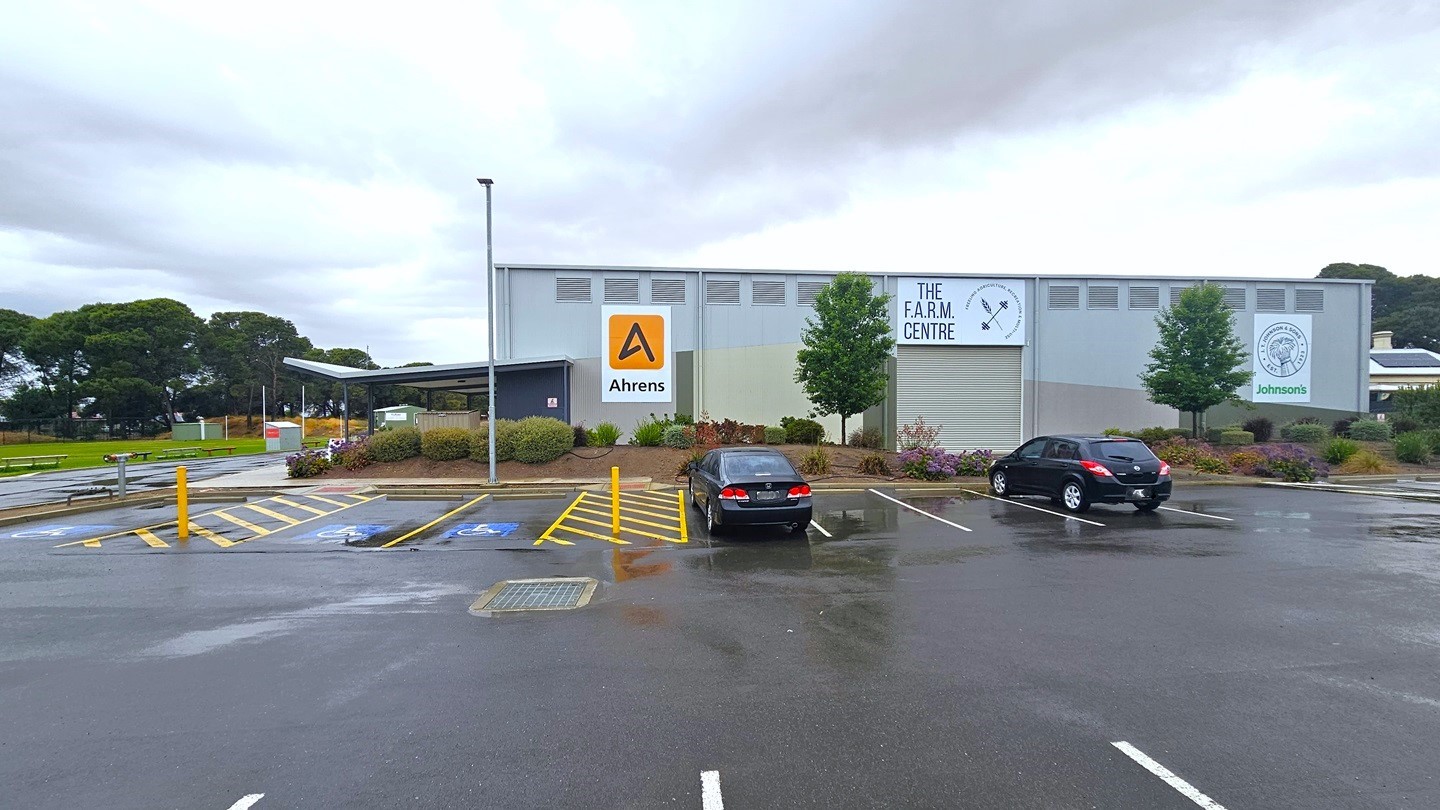 The car park is located near the building. Accessible parking is marked with blue wheelchairs, yellow diagonal lines, and bollards. Two black cars are parked. Behind the car park is a grey industrial-type building with the sign "The Farm Centre." Advert signs are displayed on the building wall. The building is surrounded by flowers, ornamental landscapes, and trees. The left section features a grassy area. The car park is located near the building. Accessible parking is marked with blue wheelchairs, yellow diagonal lines, and bollards. Two black cars are parked. Behind the car park is a grey industrial-type building with the sign "The Farm Centre." Advert signs are displayed on the building wall. The building is surrounded by flowers, ornamental landscapes, and trees. The left section features a grassy area.