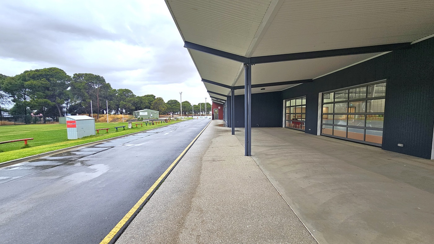 On the left is a grassy area with bench seating. The road separates the grassy area from the building on the right. The front of the building features a pitched white ceiling supported by large metal beams. On the right is a dark grey wall with large window glass. On the left is a grassy area with bench seating. The road separates the grassy area from the building on the right. The front of the building features a pitched white ceiling supported by large metal beams. On the right is a dark grey wall with large window glass.