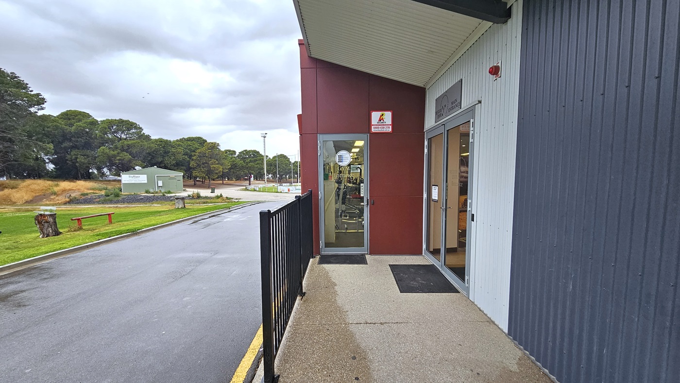 On the left is a grassy area with bench seating. A road separates the grass area from the facility building. Further ahead is a green building and a group of trees. On the right is a manual outward-opening door equipped with a non-slippery mat. Facility signage is placed on the wall. In the middle, there is a glass door that leads to the gym, with signage placed on the wall. A metal railing is positioned adjacent to the entrance. On the left is a grassy area with bench seating. A road separates the grass area from the facility building. Further ahead is a green building and a group of trees. On the right is a manual outward-opening door equipped with a non-slippery mat. Facility signage is placed on the wall. In the middle, there is a glass door that leads to the gym, with signage placed on the wall. A metal railing is positioned adjacent to the entrance.
