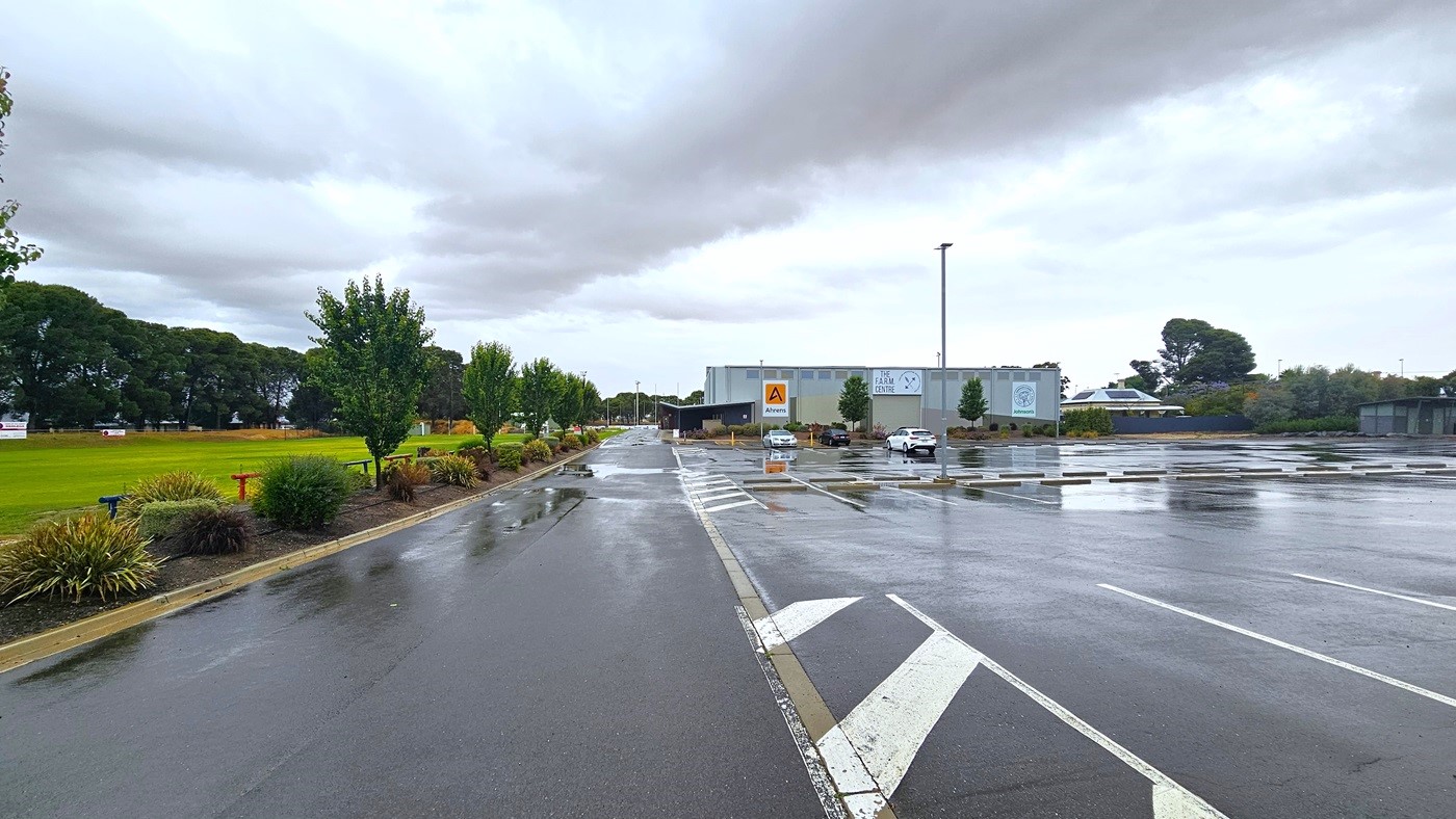 To the left is a grassy football area bordered with ornamental trees, and bench seatings are visible. On the right is an industrial building painted in grey, bearing the facility signage and some adverts. The car park is located at the side of the building, with three cars currently parked. To the left is a grassy football area bordered with ornamental trees, and bench seatings are visible. On the right is an industrial building painted in grey, bearing the facility signage and some adverts. The car park is located at the side of the building, with three cars currently parked.
