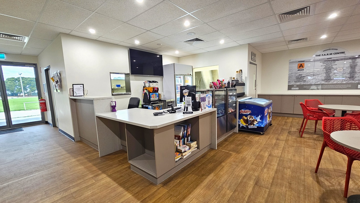 The reception desk, with a cream desk, is in the middle. A flat-screen TV is on the wall, and books are on the shelves underneath. Beside it is a display rack showcasing the cafe's items. Movable red chairs and white tables are on the right. A large poster on the wall has information. The timber floor is made of timber and the ceiling is white. To the right is a lobby leading to other facilities. On the left is a glass door with a fire extinguisher and pamphlet holder on the wall. The reception desk, with a cream desk, is in the middle. A flat-screen TV is on the wall, and books are on the shelves underneath. Beside it is a display rack showcasing the cafe's items. Movable red chairs and white tables are on the right. A large poster on the wall has information. The timber floor is made of timber and the ceiling is white. To the right is a lobby leading to other facilities. On the left is a glass door with a fire extinguisher and pamphlet holder on the wall.